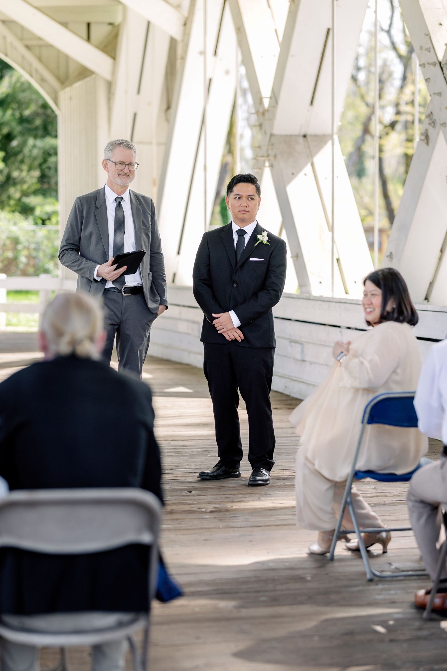 Groom in black suit stands at wedding altar with officiant holding clipboard at outdoor covered bridge ceremony.