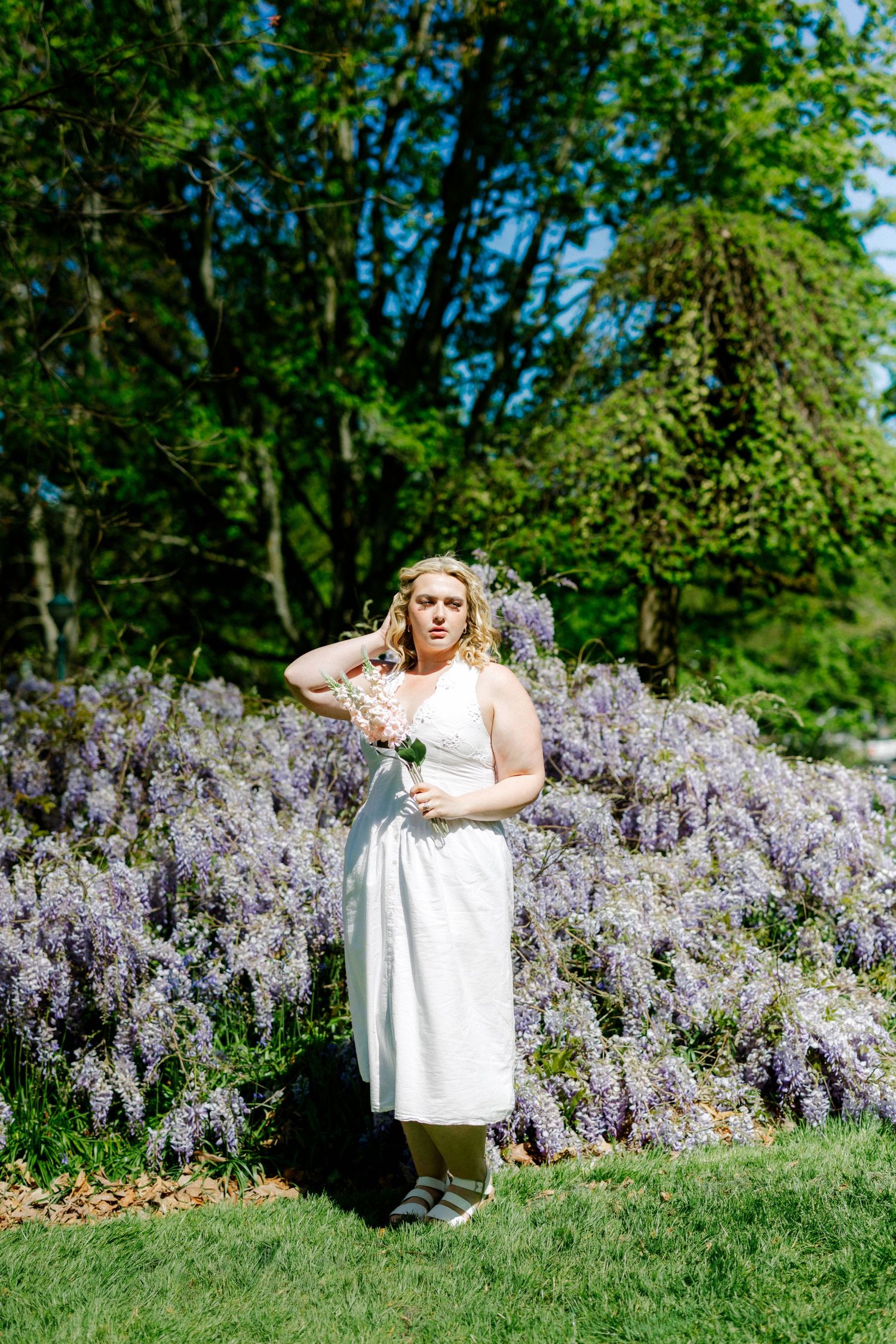 Woman in white dress holding flowers stands before purple wisteria blooms in a sunny garden.