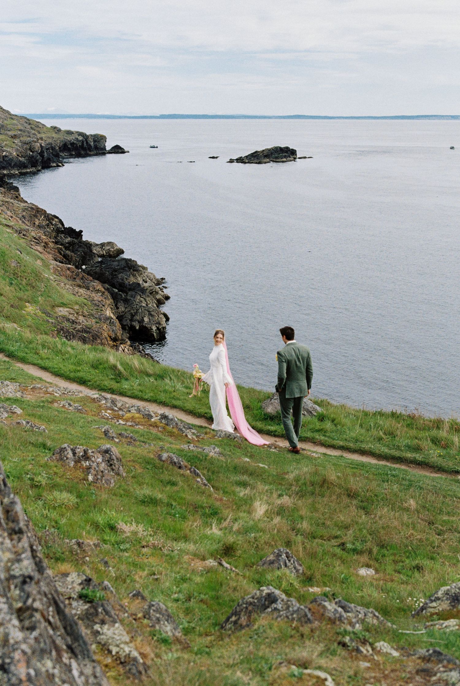 Bride in white gown with pink train and groom in green suit walk coastal cliff path above rocky shoreline.