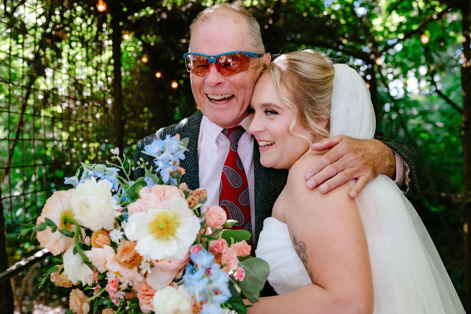 Smiling bride hugs older man wearing red sunglasses while holding colorful wedding bouquet outdoors.