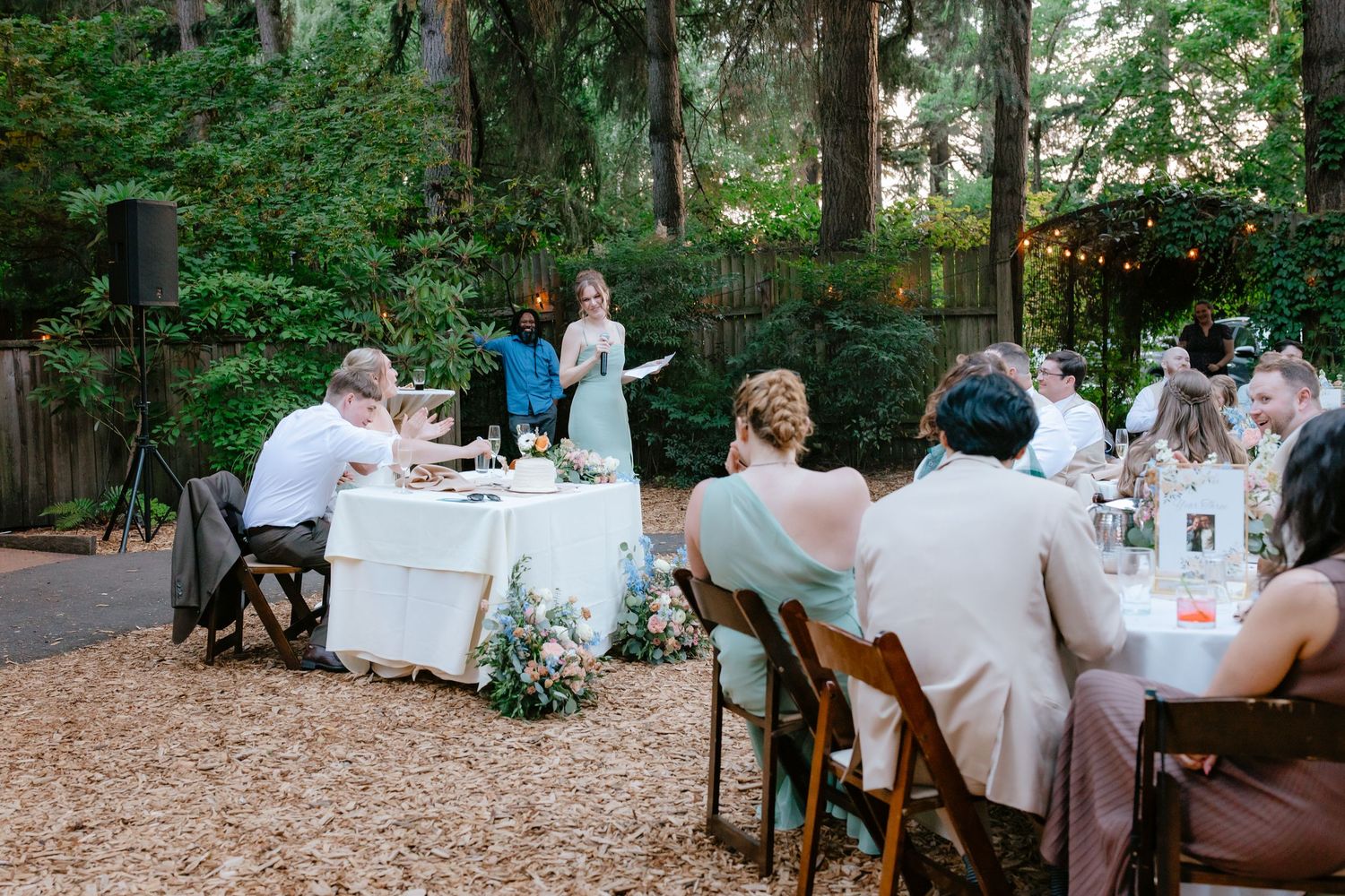 Wedding guests seated at outdoor reception dinner tables surrounded by trees and string lights.
