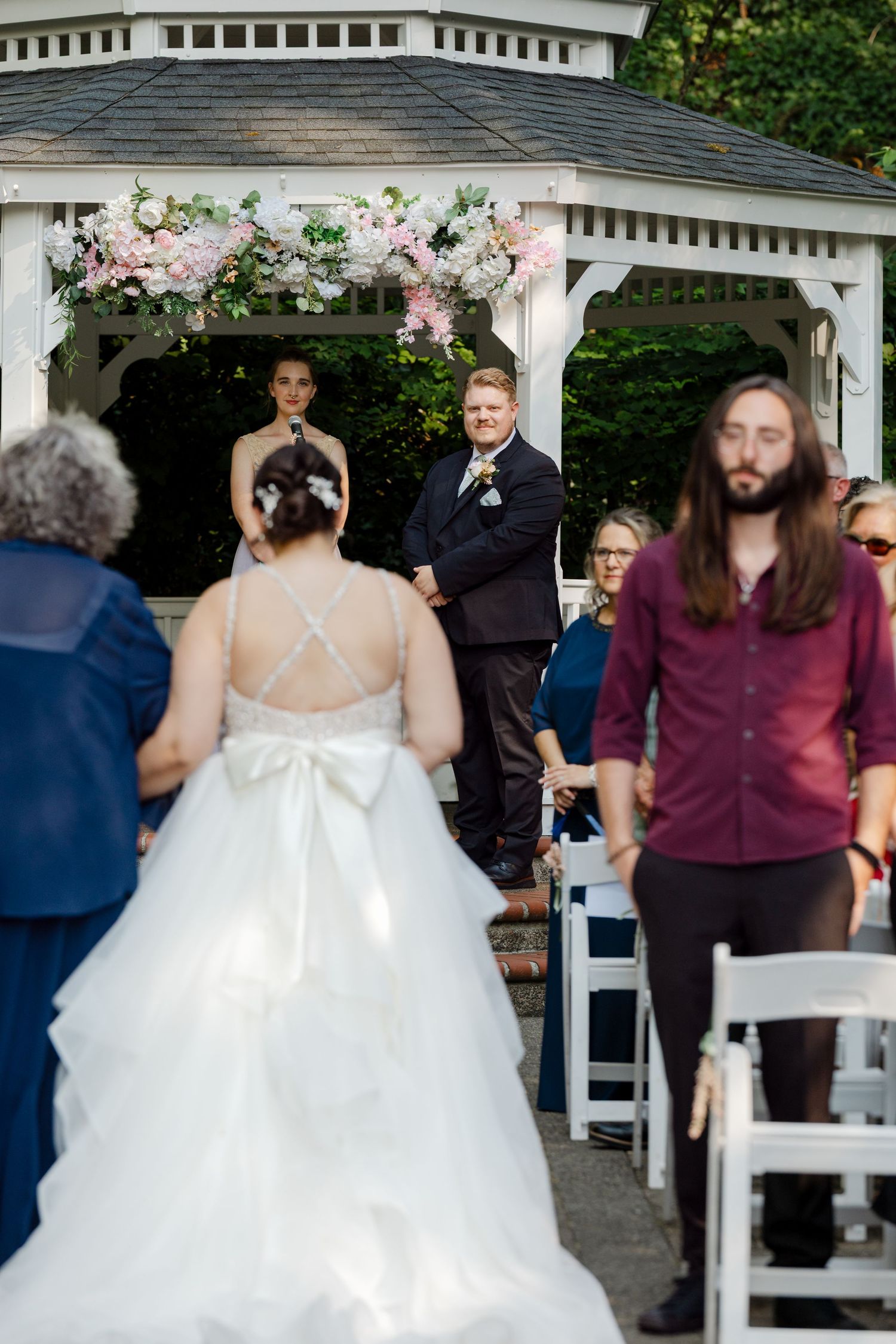 Bride walking down the aisle toward the groom standing at a floral-decorated gazebo during the ceremony.