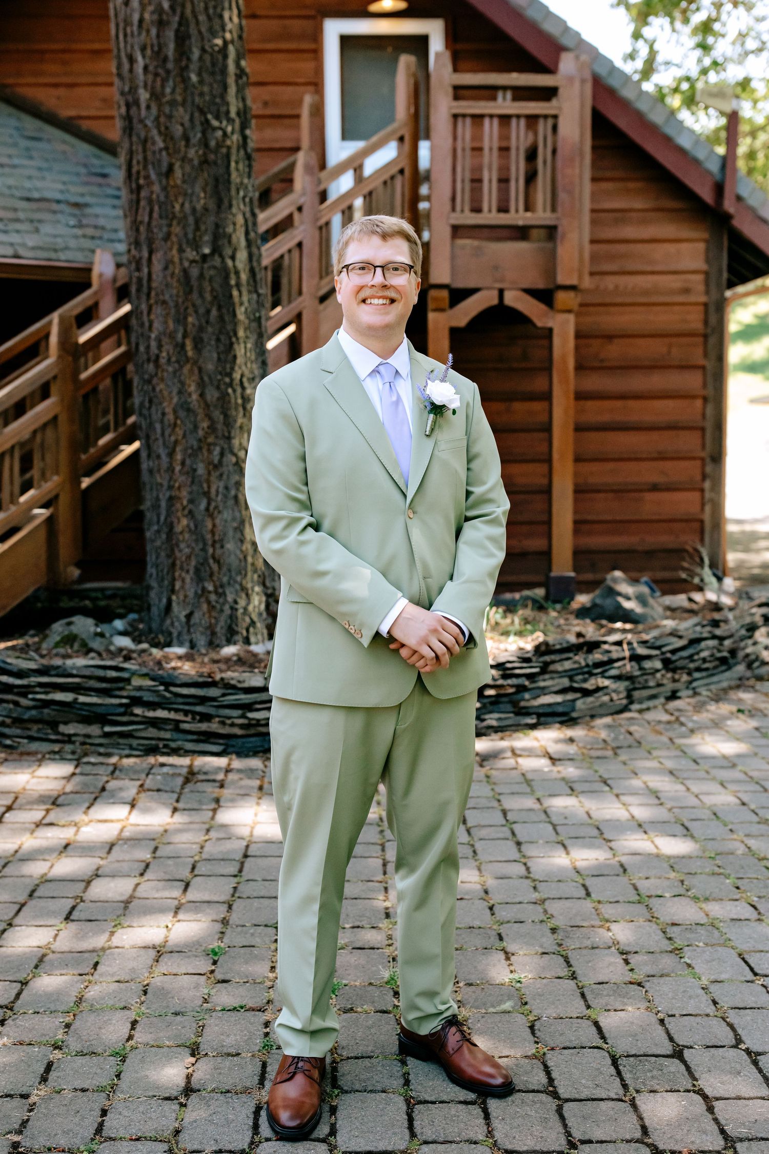 Smiling groom in sage green suit with boutonniere stands before rustic wooden cabin on wedding day.