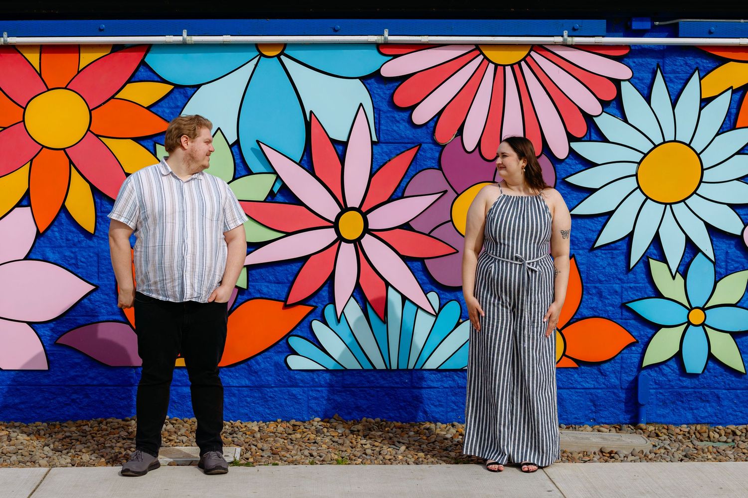 Two people pose against a vibrant floral mural with colorful daisies and geometric patterns.