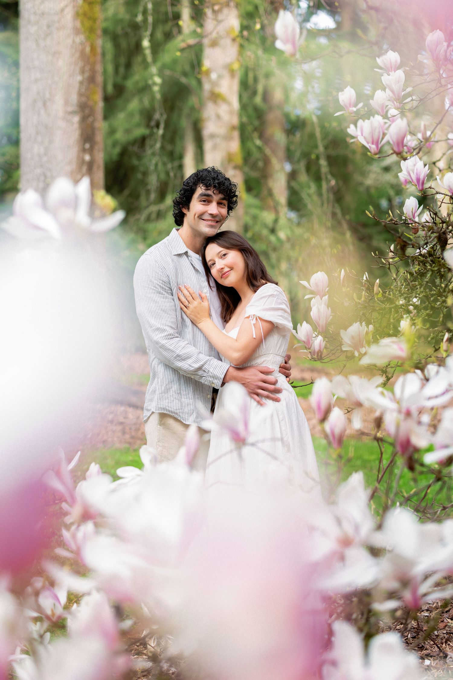 A romantic couple embraces among blooming pink magnolia flowers in a dreamy spring garden setting.