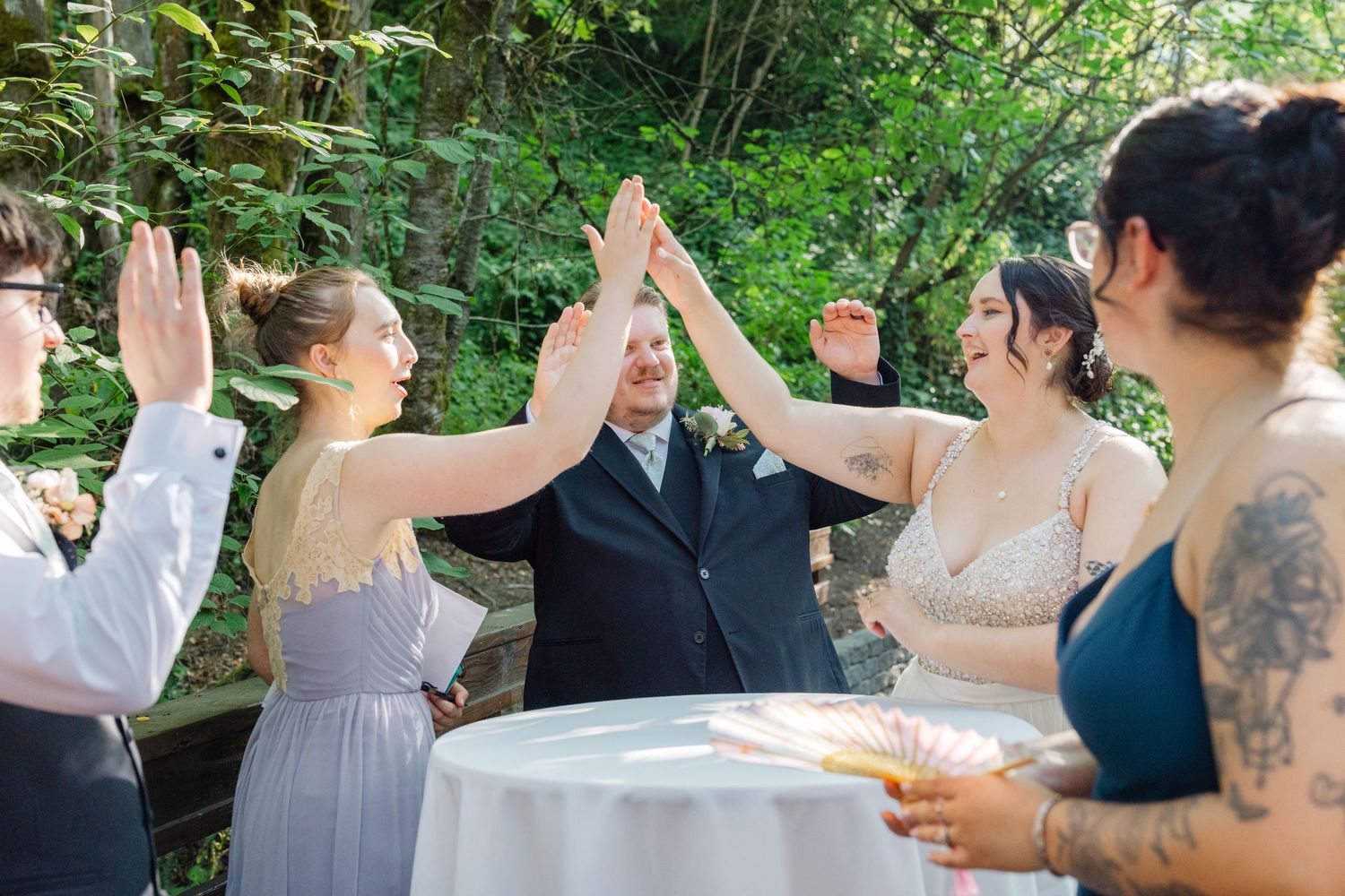 Bride, groom, and wedding party celebrate with a group high-five at an outdoor garden reception.