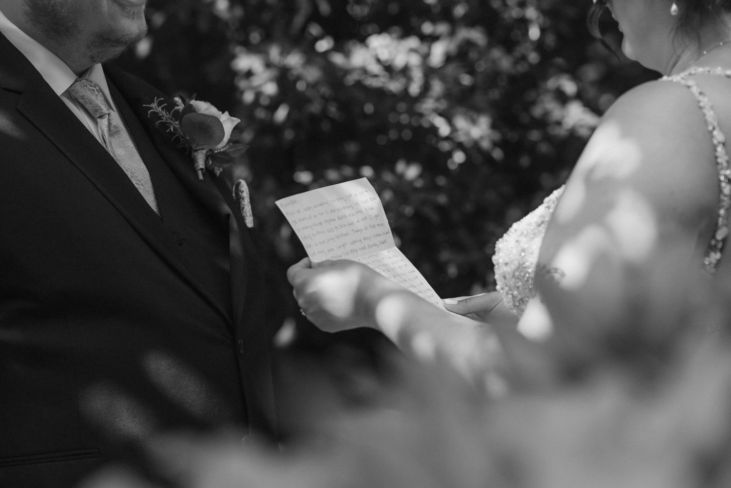 Black and white wedding photo of bride reading vows from paper to groom during outdoor ceremony.
