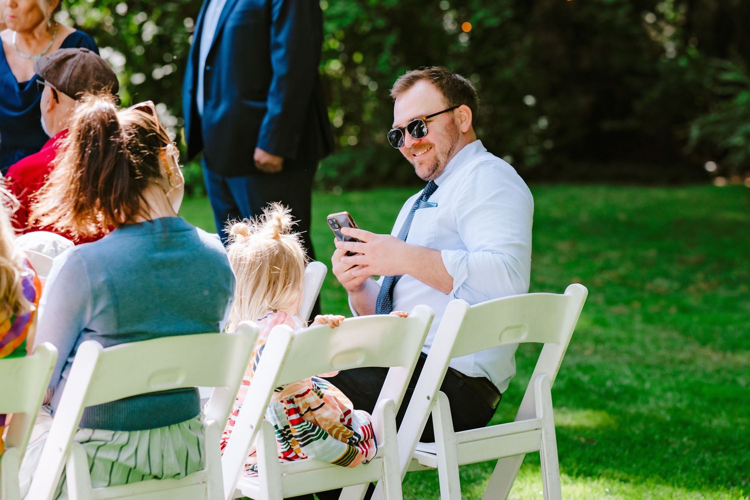 A man in sunglasses checks his phone while seated at an outdoor wedding ceremony.