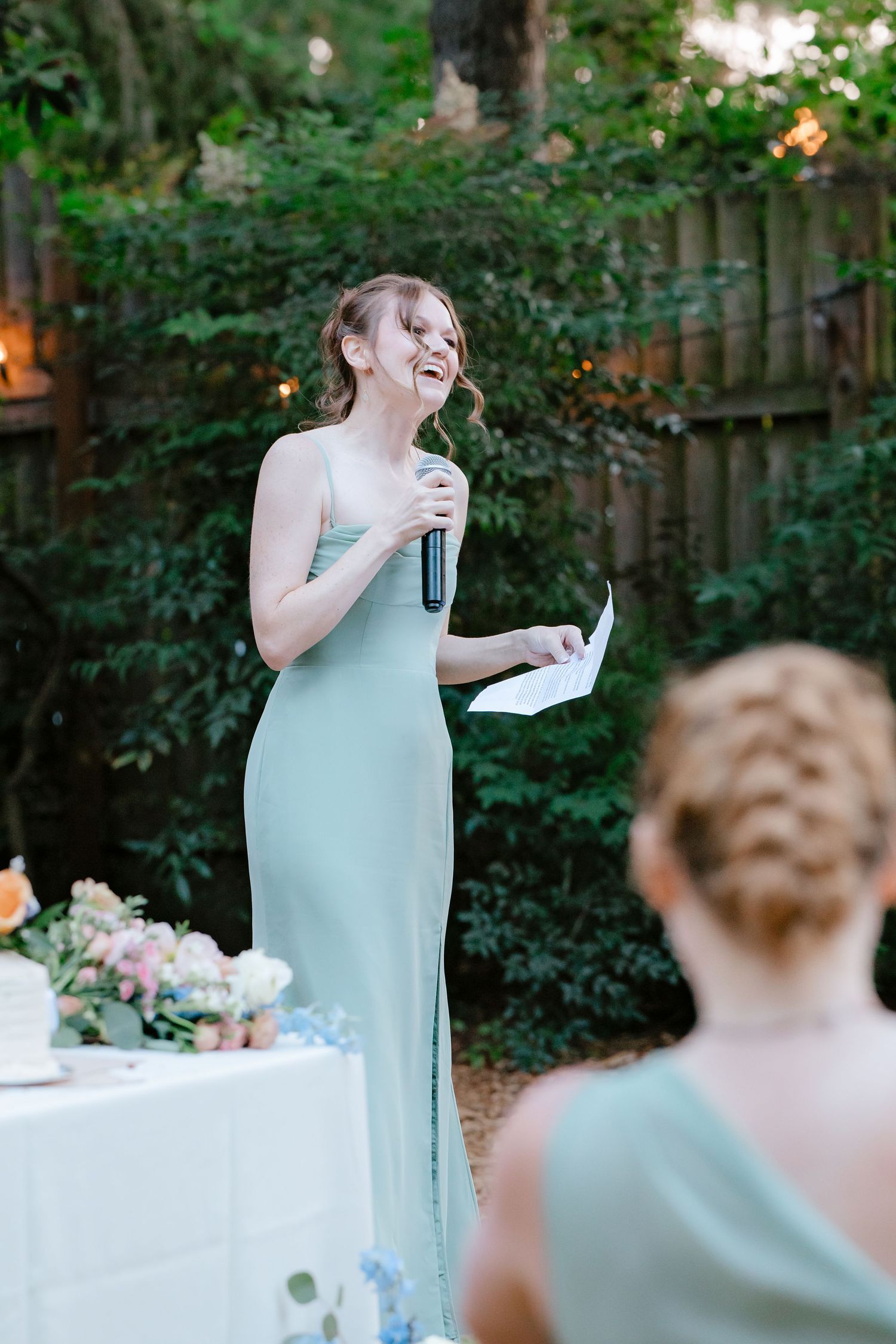 Bridesmaid in sage green gown gives speech at outdoor wedding reception.