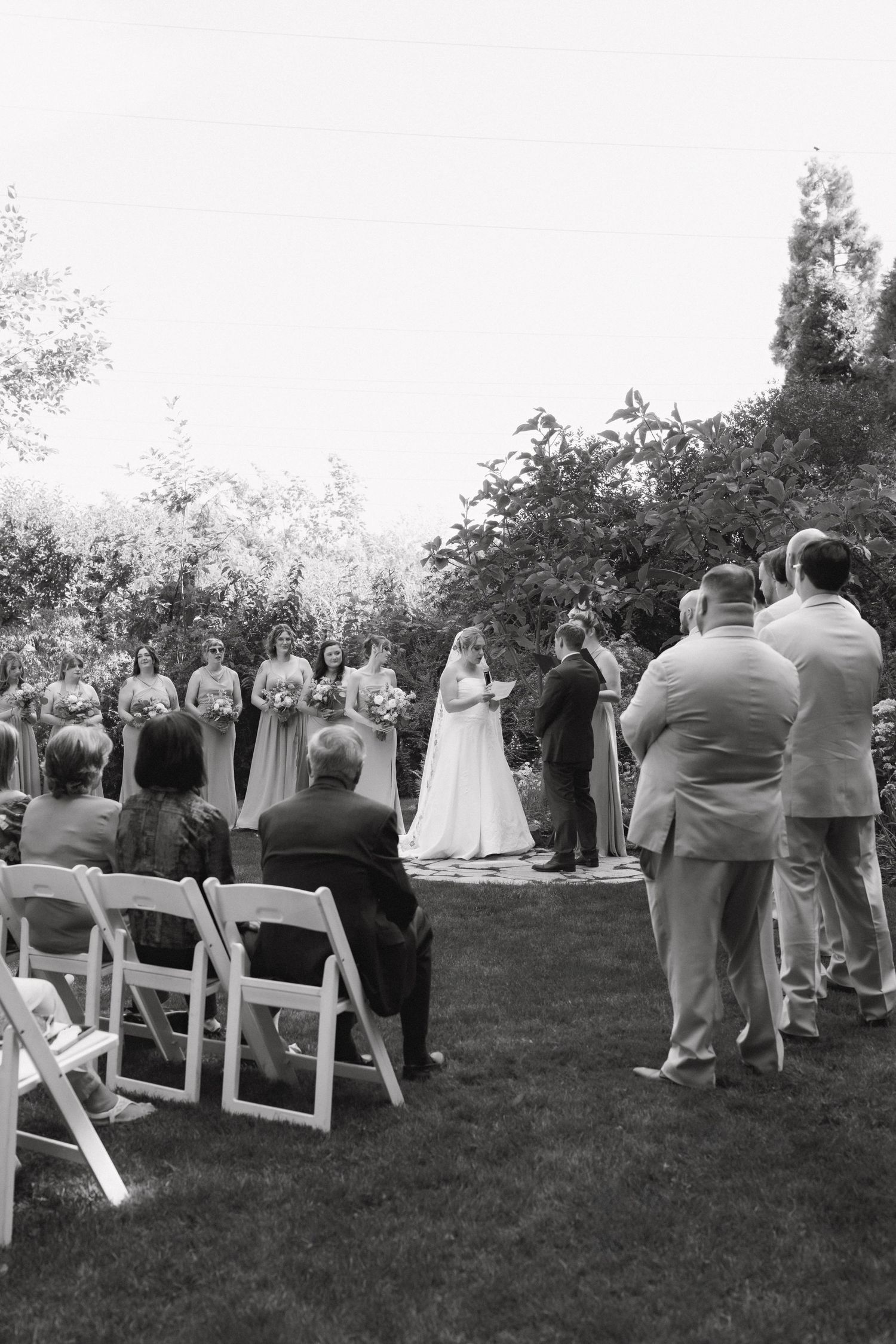 Black and white outdoor wedding ceremony with bride, groom, bridesmaids, and seated guests on a lush garden lawn.