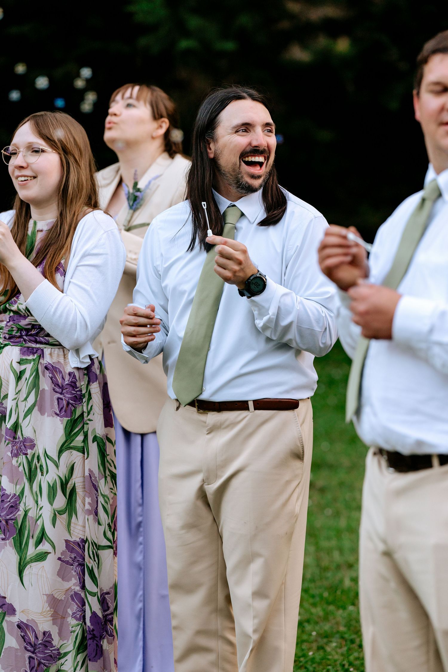 Joyful wedding guest in white shirt and green tie laughs during outdoor wedding reception celebration.