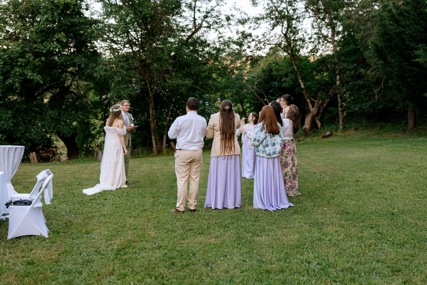 Wedding party stands in a circle on green lawn at dusk surrounded by lush trees during outdoor ceremony.