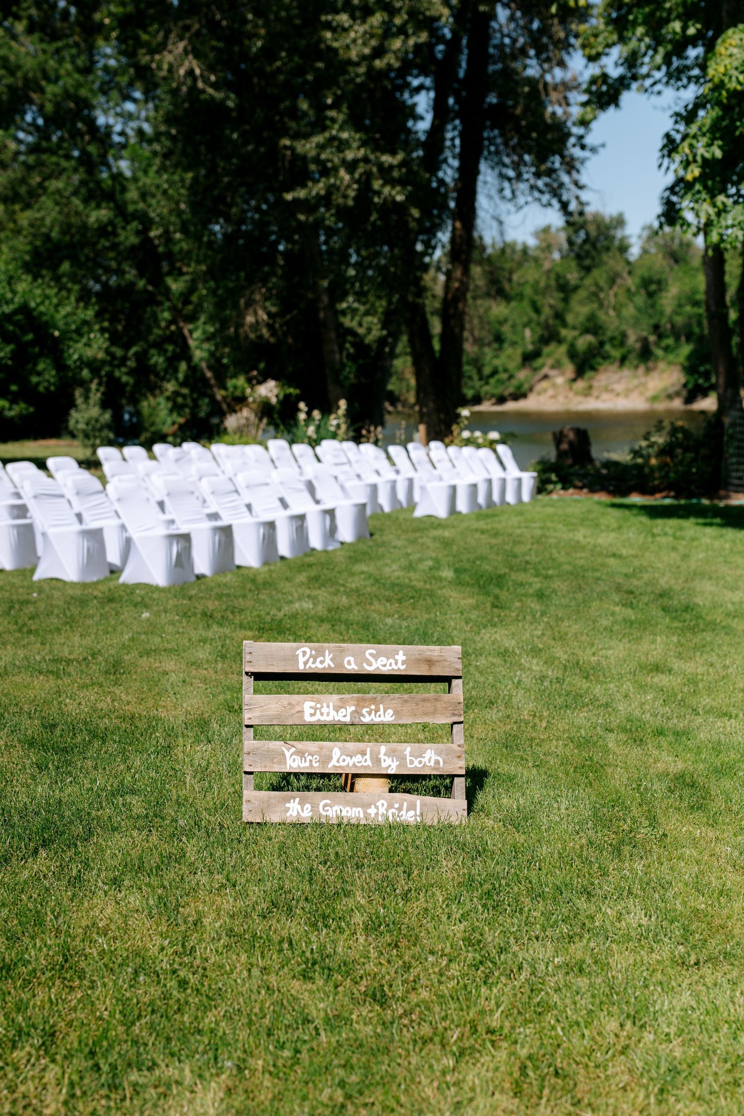 Rustic wooden pallet wedding sign reading Pick a Seat Either Side on a green lawn with white chairs arranged for an outdoor ceremony.