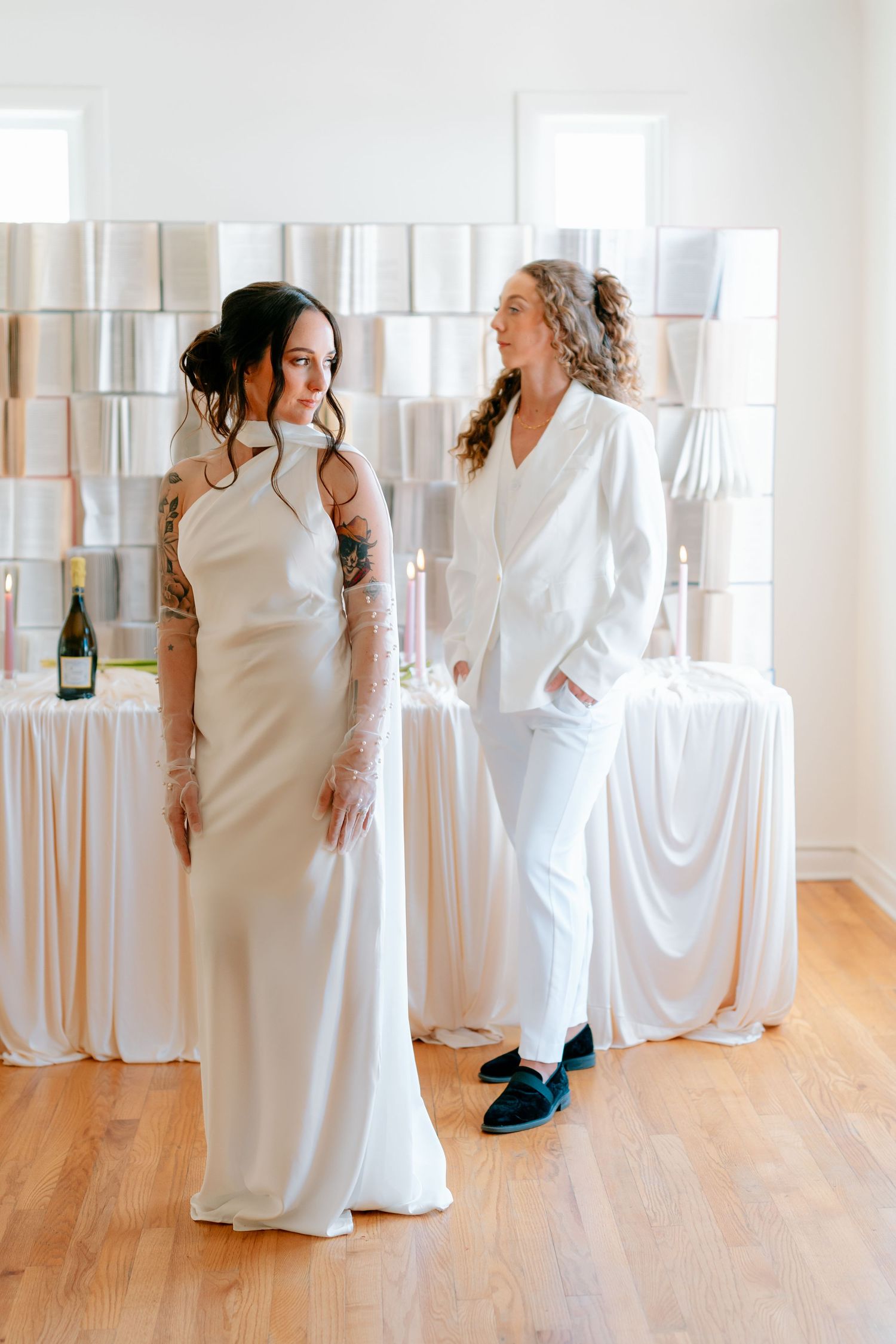 Two brides in white wedding attire, one in a satin gown and one in a suit, standing in an elegant bright venue.