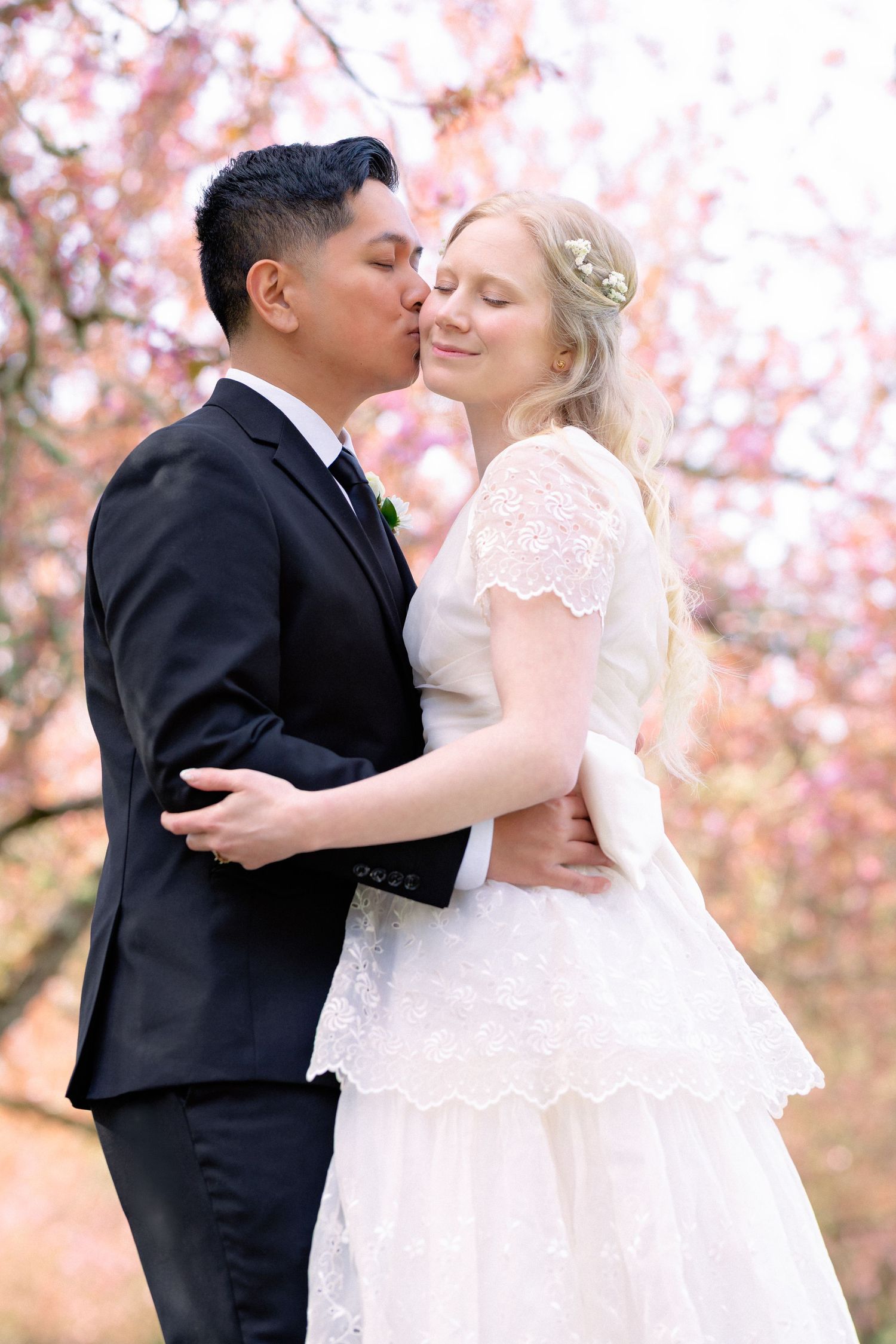 A groom in a black suit tenderly kisses his blonde bride on the cheek beneath pink cherry blossom trees.