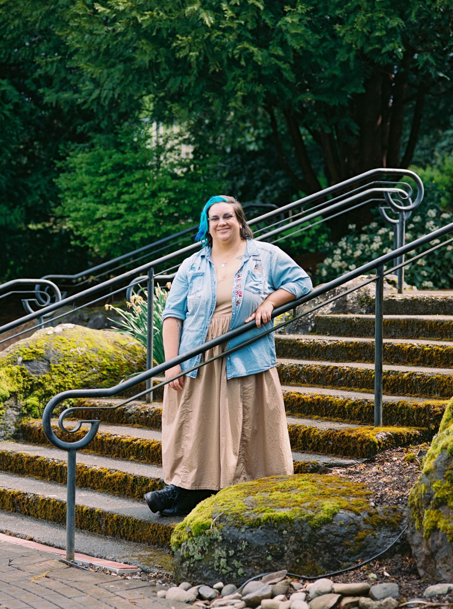 Smiling woman with blue hair wearing a tan dress and denim jacket stands on moss-covered stone steps outdoors.