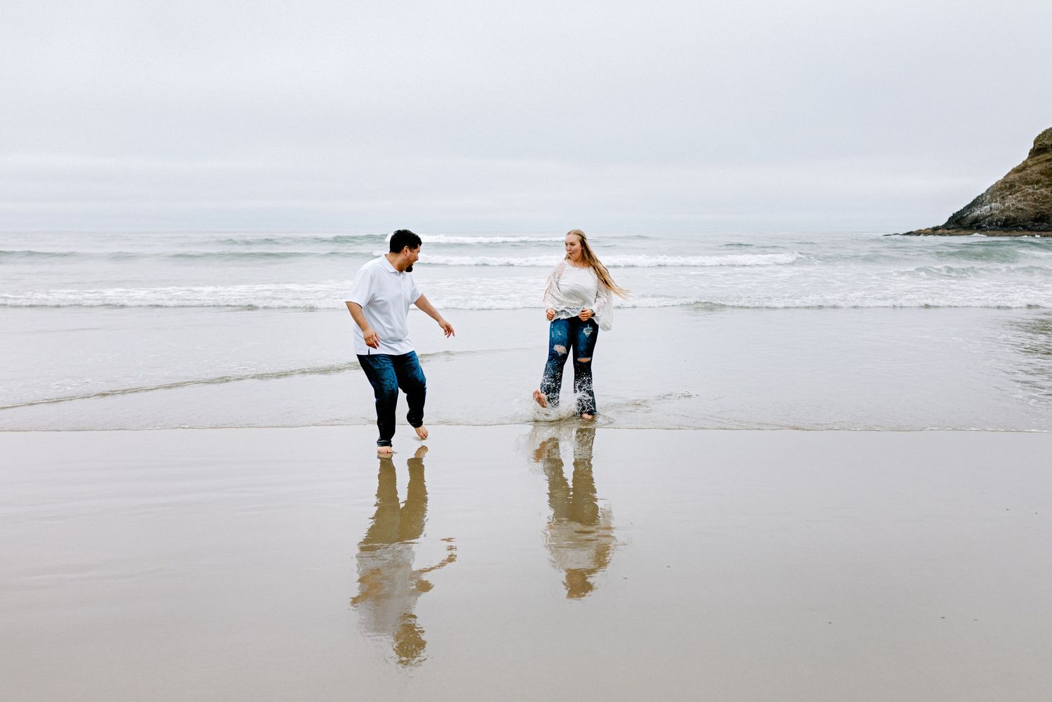 Couple playfully splashing in ocean waves on a reflective wet sandy beach under an overcast sky.