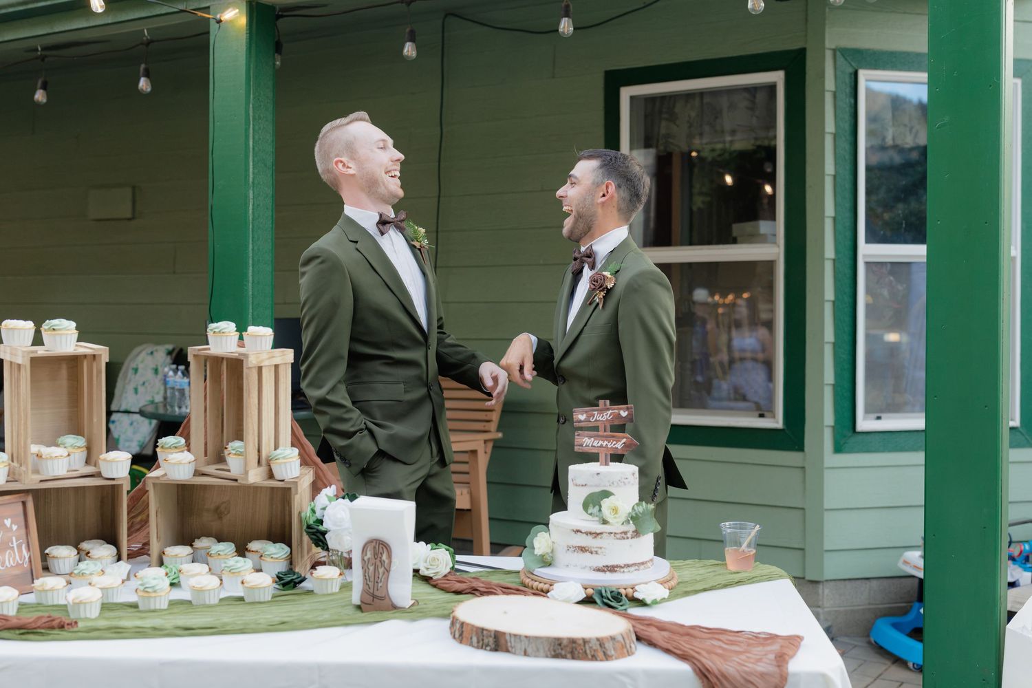 Two grooms in olive green suits share a laugh beside a dessert table at their outdoor wedding reception.