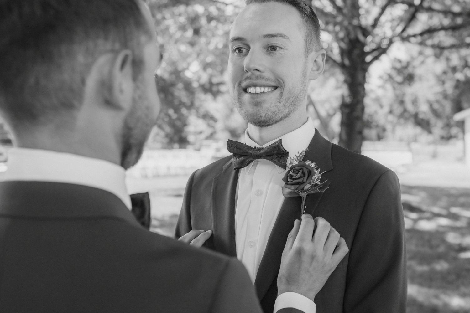 Two grooms in tuxedos share a tender moment outdoors, one adjusting the other's boutonniere at their wedding.