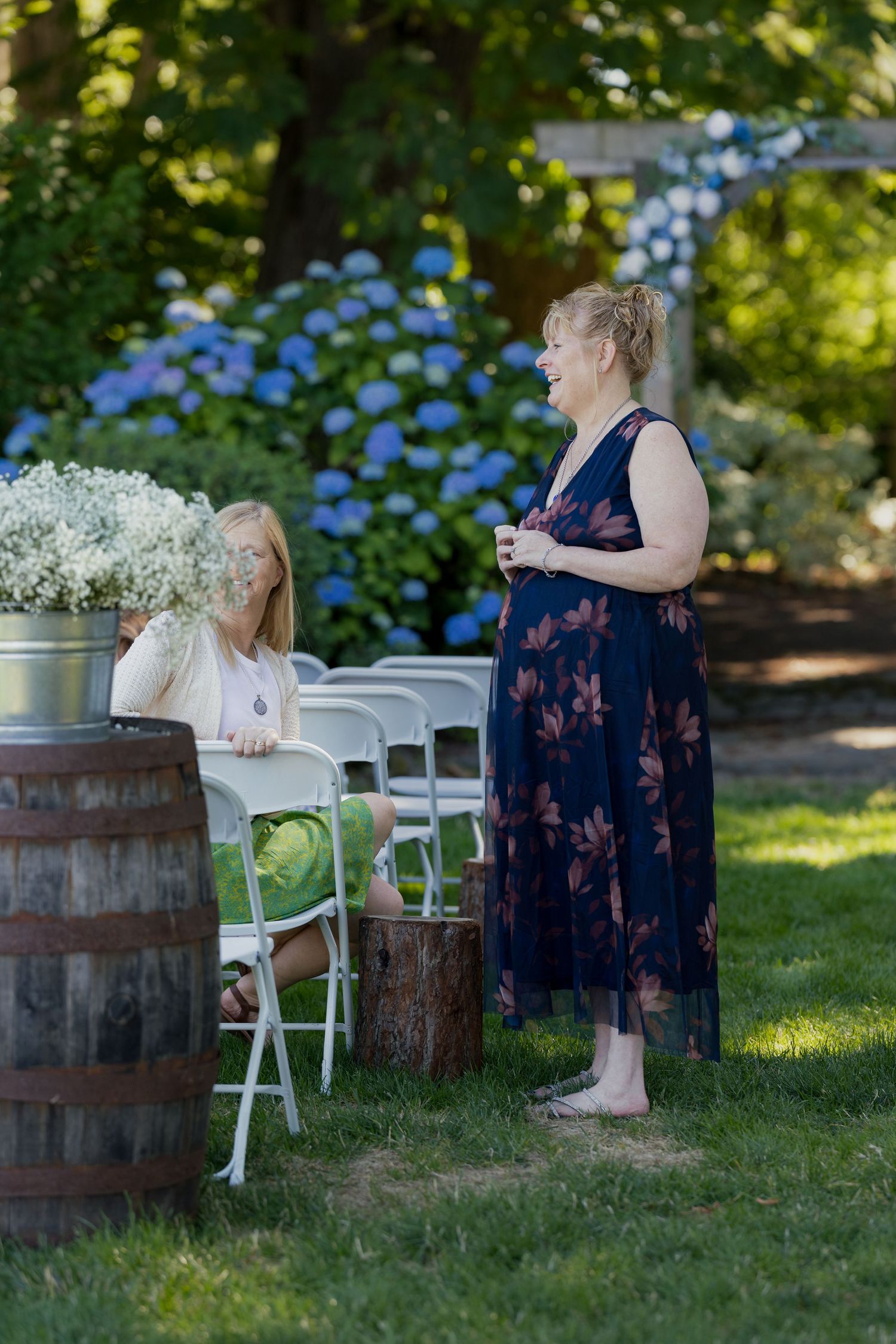 Two women conversing outdoors at a garden wedding with hydrangeas, white chairs, and a rustic wooden barrel decoration.