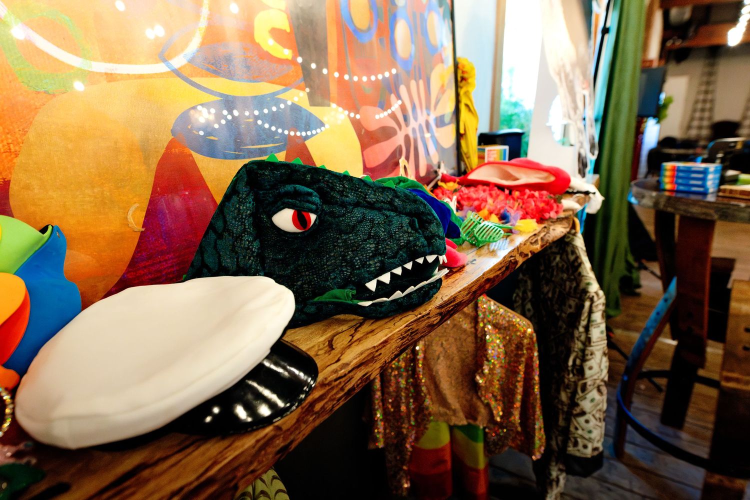 Colorful party table displays dinosaur costume head, frisbee, and festive accessories against vibrant mural backdrop.