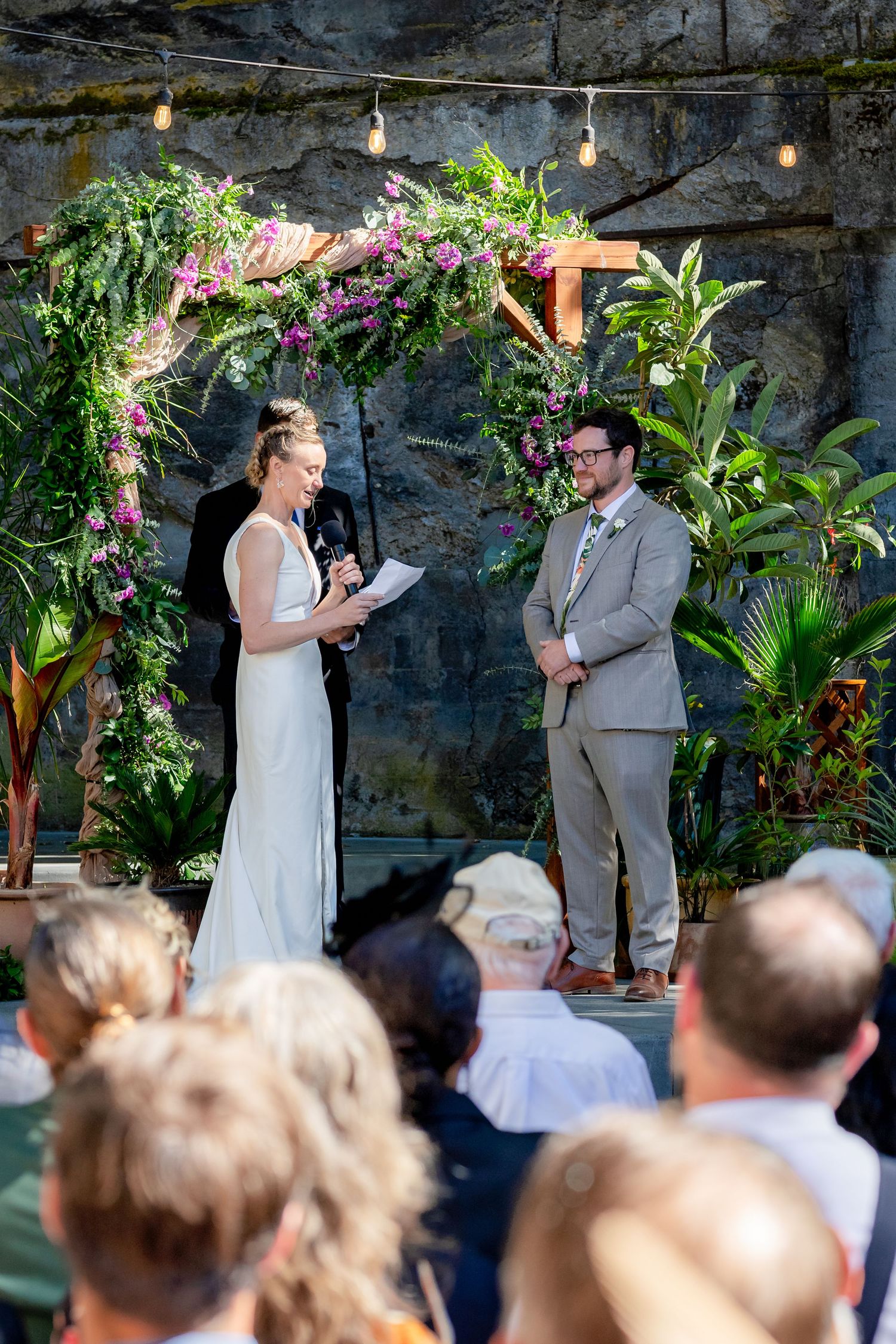 Bride reads vows at outdoor wedding ceremony under floral arch with purple orchids as groom listens before seated guests.