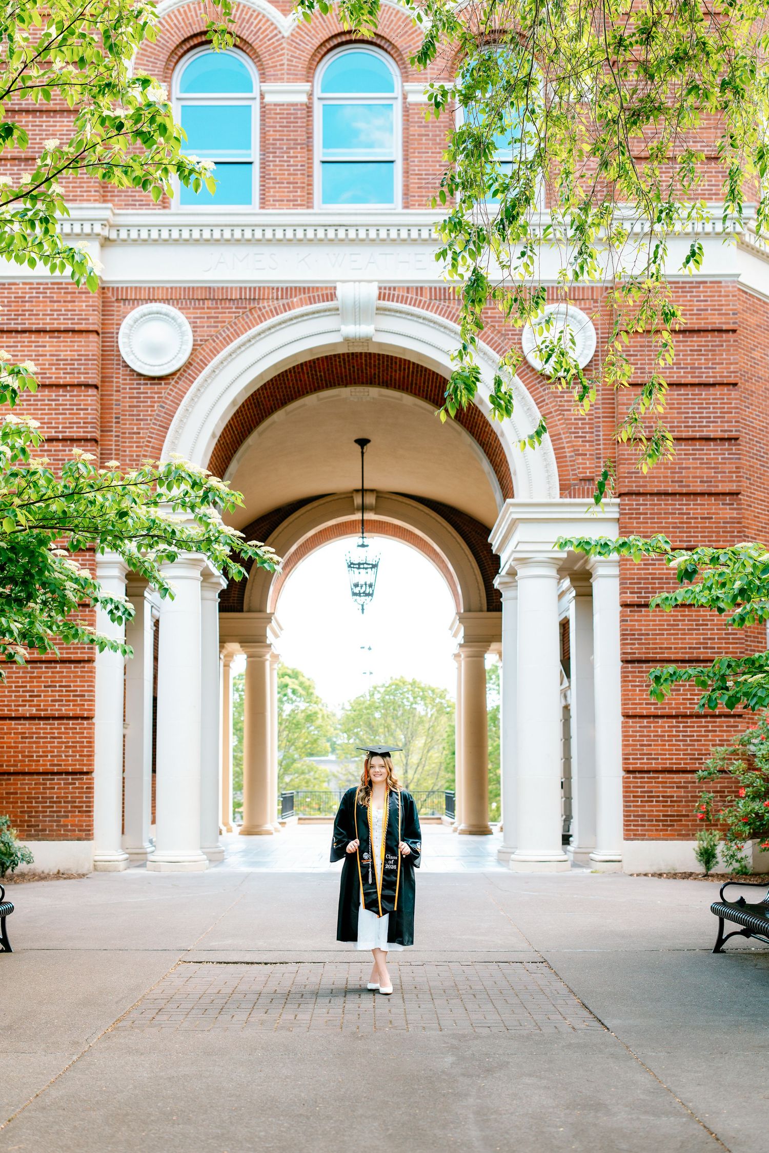 A graduate in black cap and gown stands under a brick archway at Oregon State University on a sunny day.