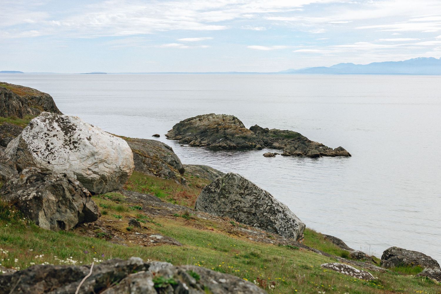 Rocky coastal shoreline with lichen-covered boulders overlooking calm ocean waters and distant mountains under overcast skies.