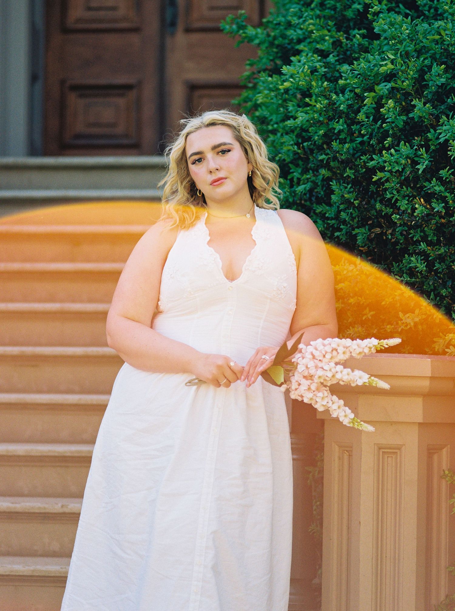 Plus-size blonde woman in white scalloped dress holding pink flowers on sunlit stone steps.