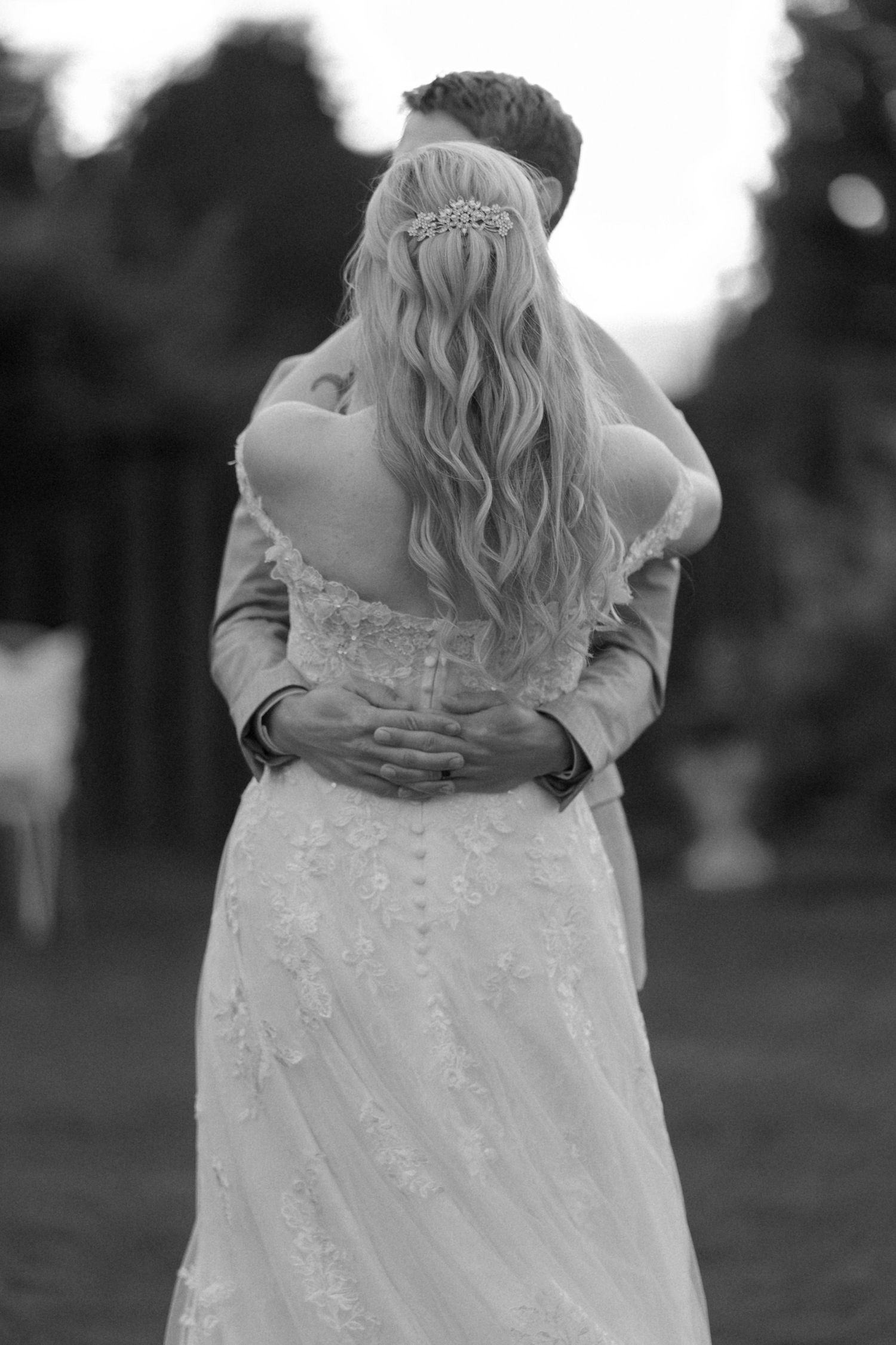 Black and white photo showing back view of a lace wedding dress during an outdoor celebration.