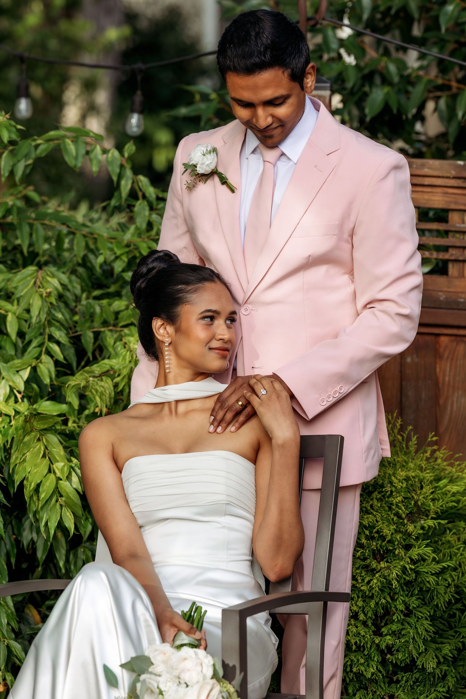Couple shares tender moments on wooden bench in garden, wearing white dress and pink suit.