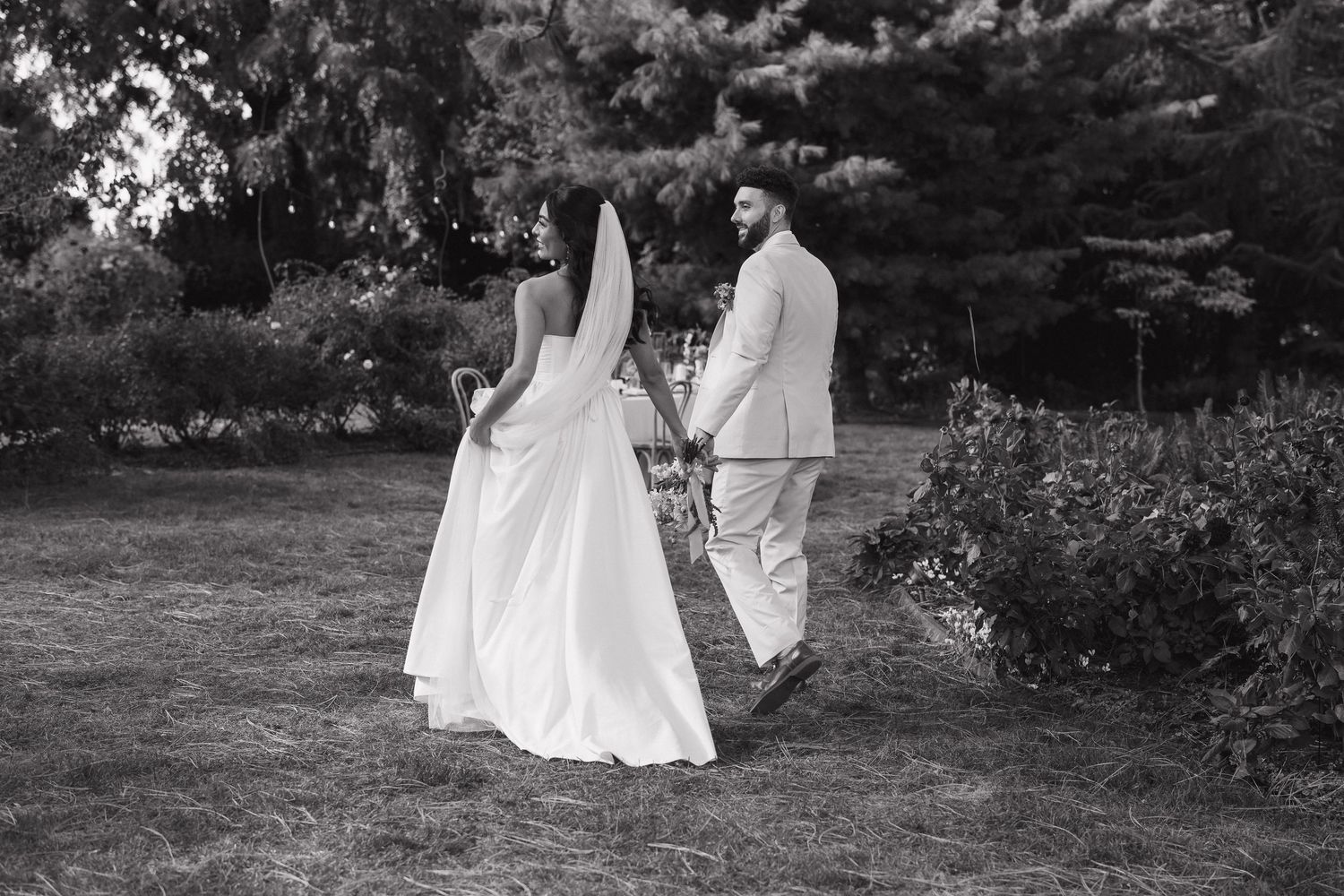 Newlywed couple walks through a garden path holding hands in elegant white wedding attire.