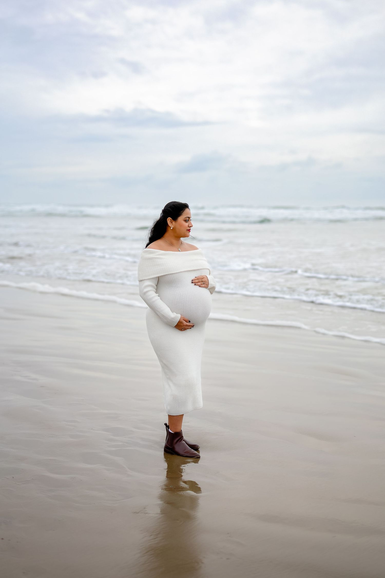 A person in a white dress stands at the edge of the ocean shore as waves roll in on a cloudy day.