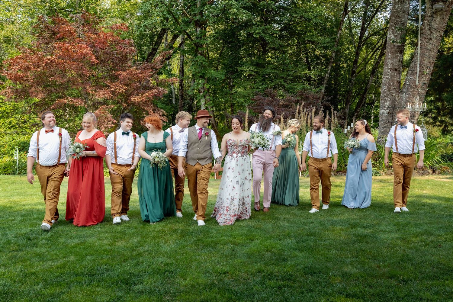 Wedding party members in colorful attire posed on grass with trees in the background.