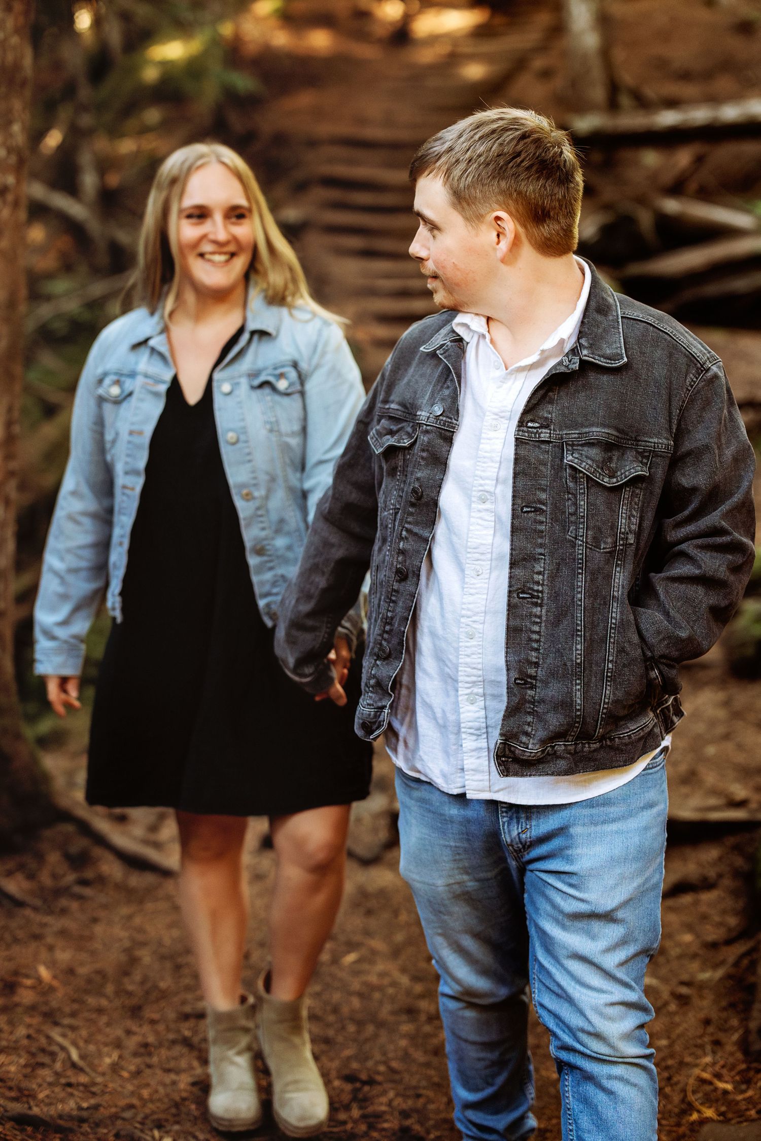 Casual couple in denim jackets walking through a wooded forest setting.