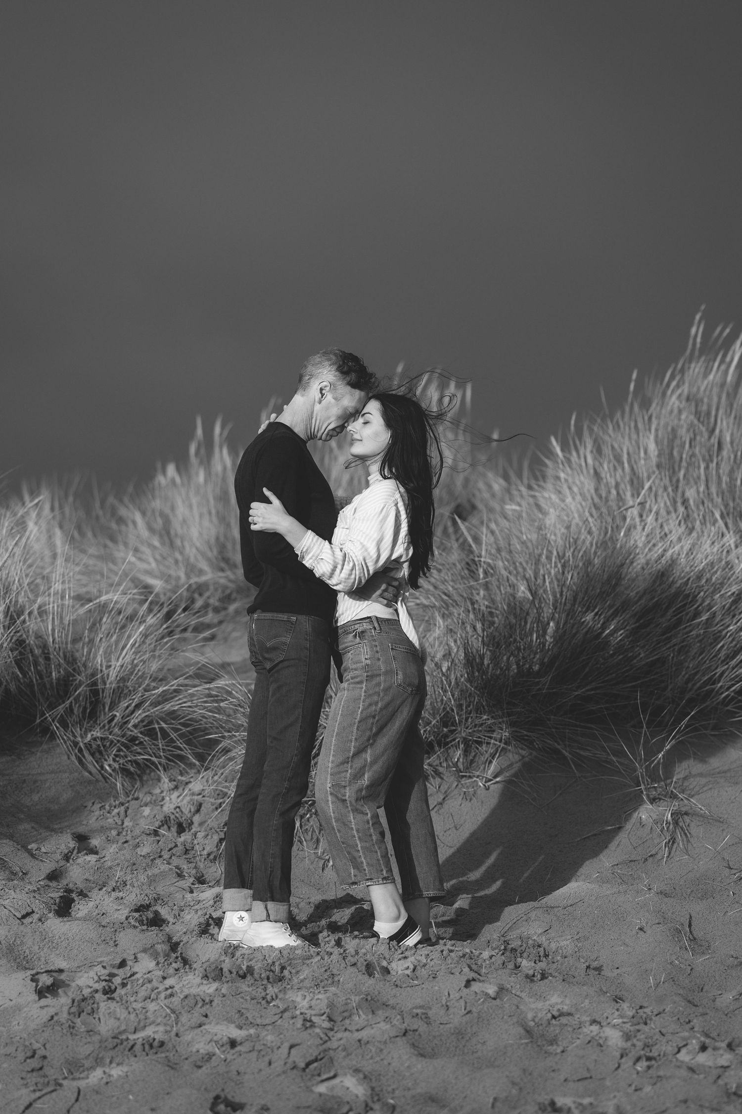 A series of black and white photographs showing two figures embracing on a sandy beach with tall grass in the background.