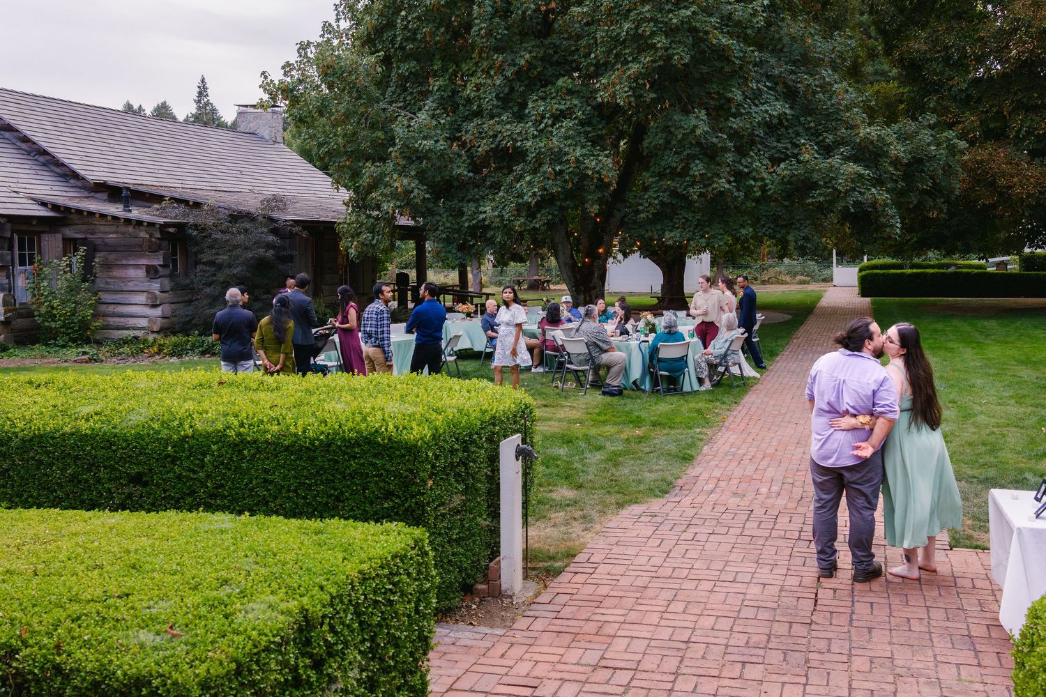 Brick pathway leads through garden with people walking and gathering near historic buildings.