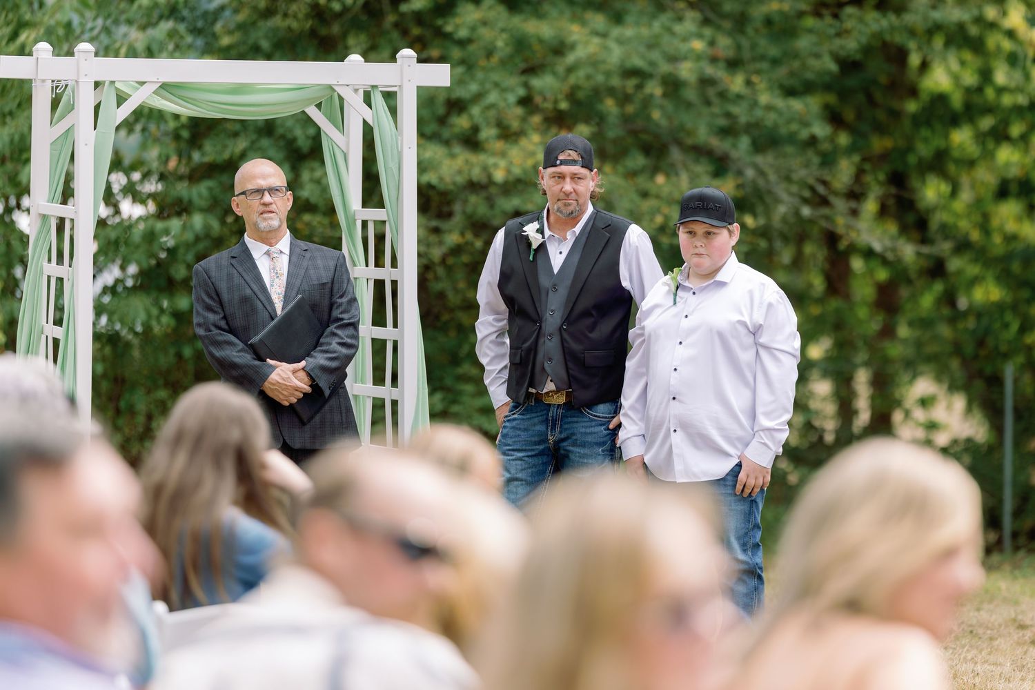 Three people stand under a white wedding arch with guests seated in the foreground during an outdoor ceremony.