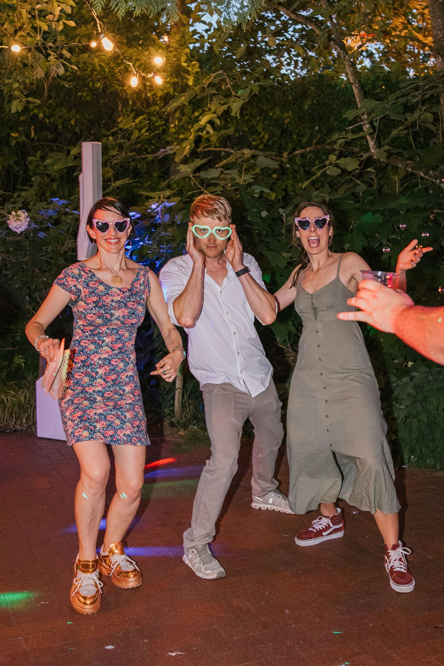 People dance in a line at an outdoor evening party with twinkling lights and trees in the background.