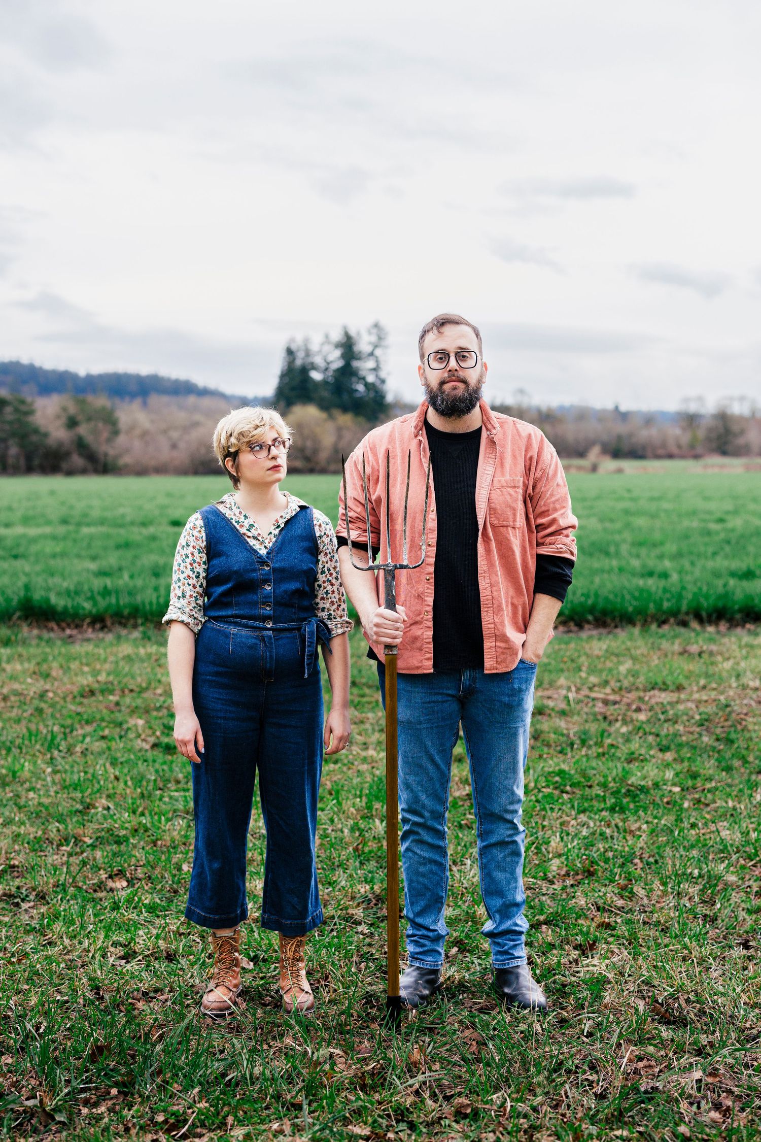 Two figures stand in a grassy field wearing casual attire with mountains and trees visible in the distance.