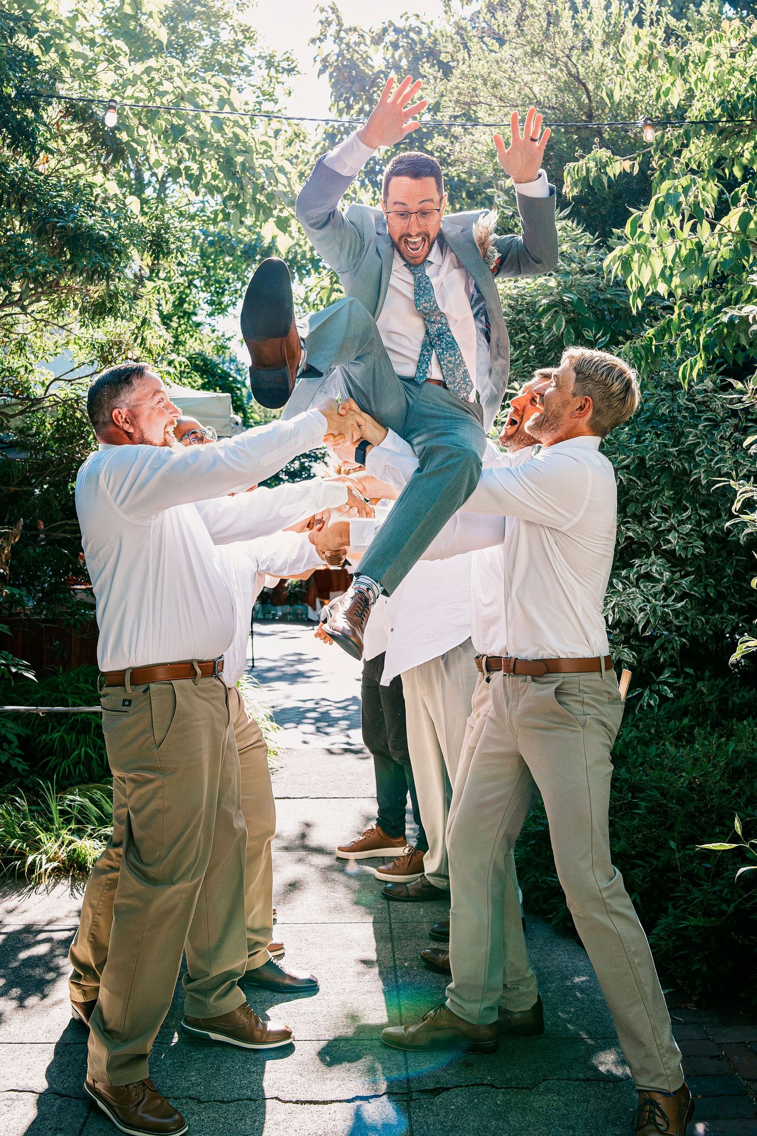 A sequence of playful groomsmen jumping and posing together in khaki pants and white shirts in an outdoor garden setting.