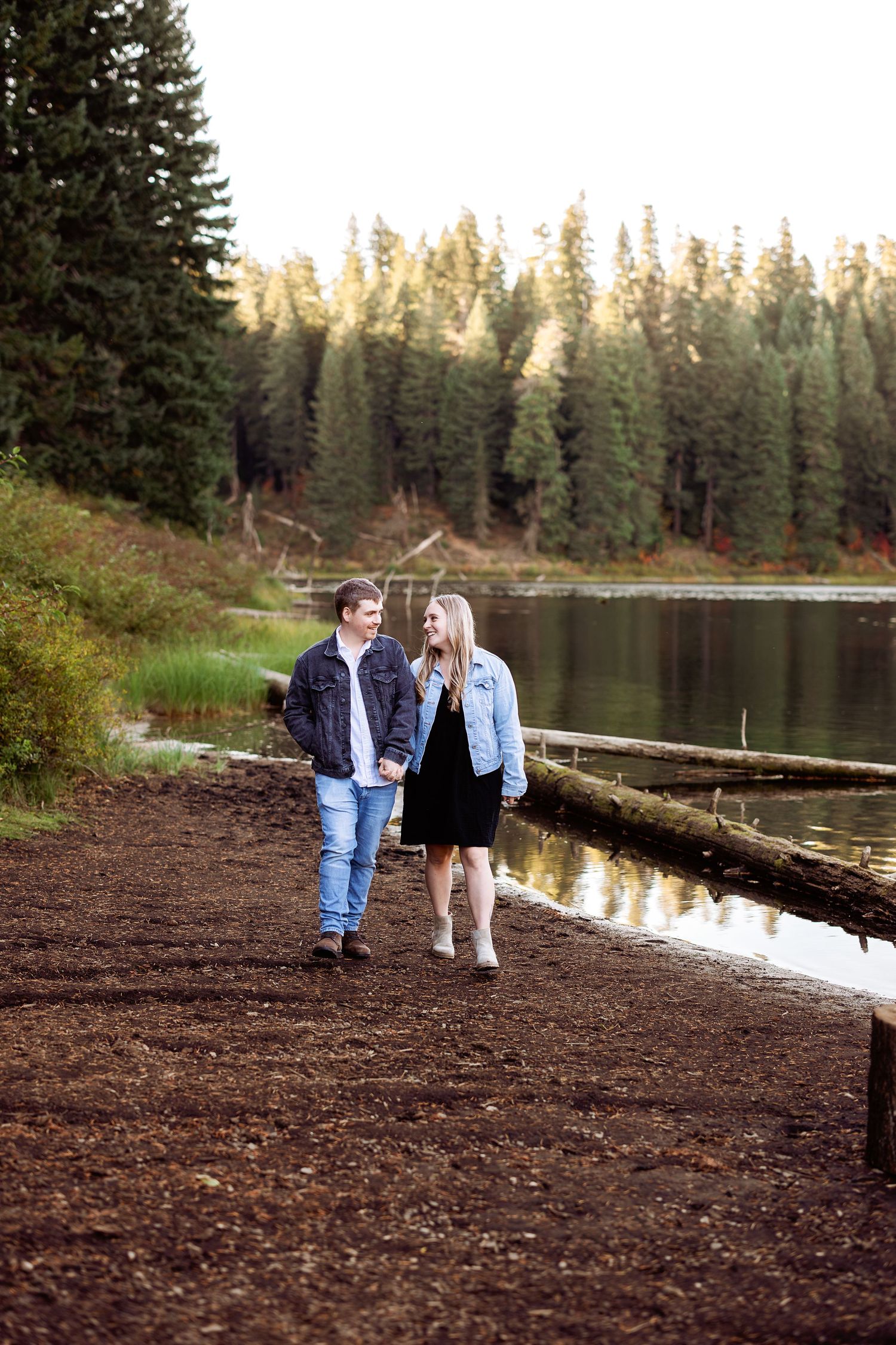 A couple sitting together on a log by a serene lake surrounded by pine trees at sunset.