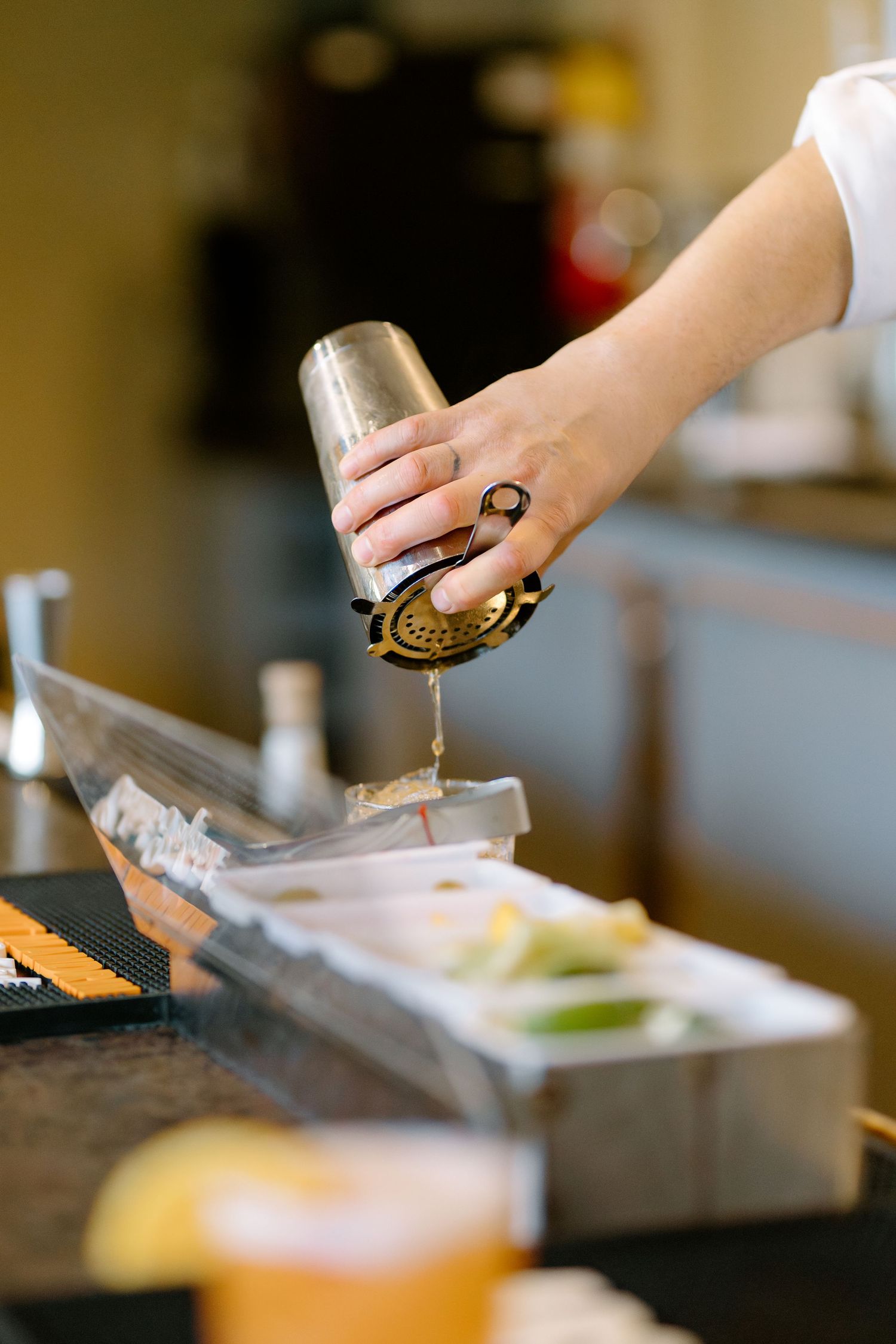 Bartender straining a cocktail through a gold strainer into a bar setup with fresh citrus garnishes.
