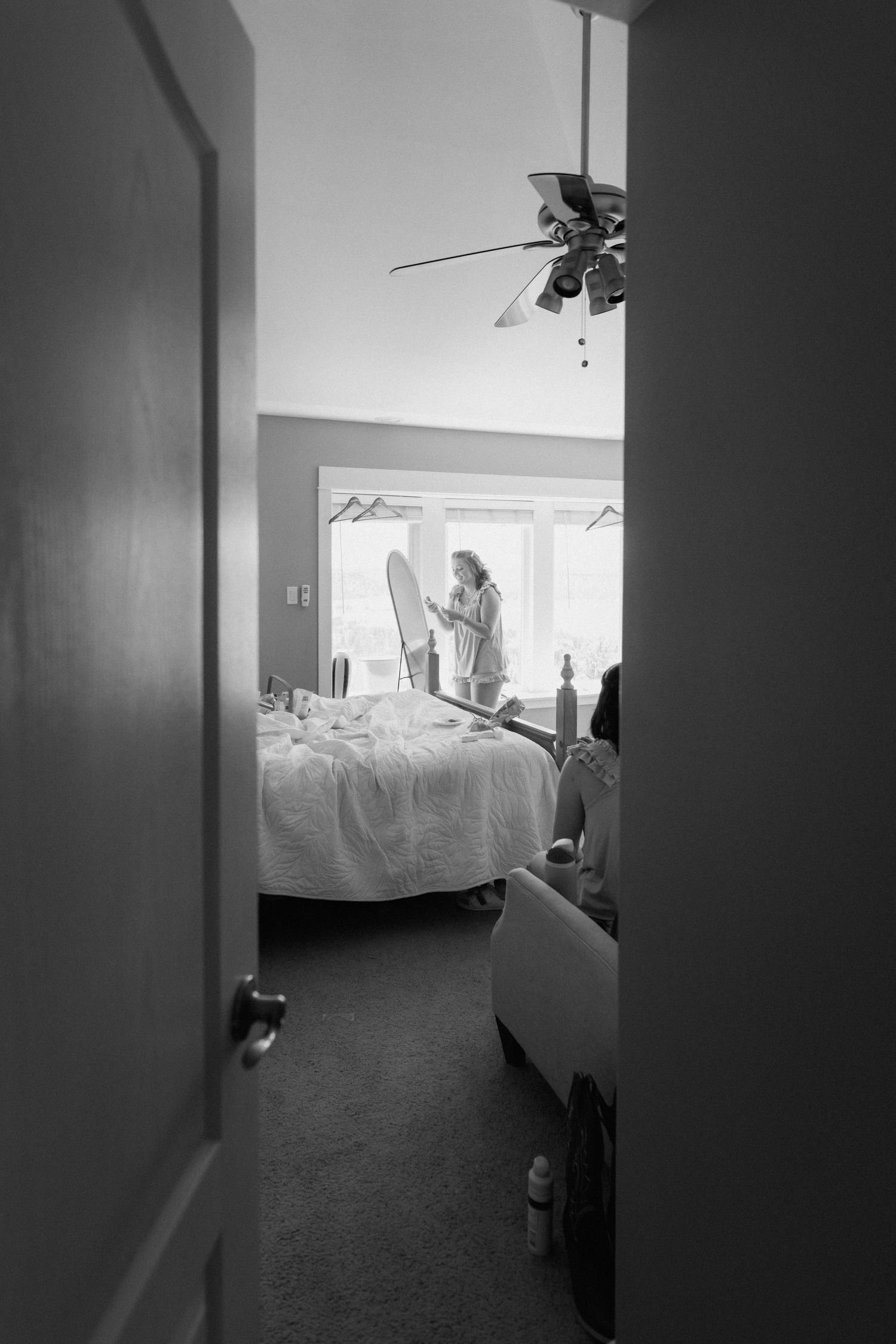 Black and white photo of a bride getting ready in a bedroom, viewed through a doorway with a ceiling fan above.