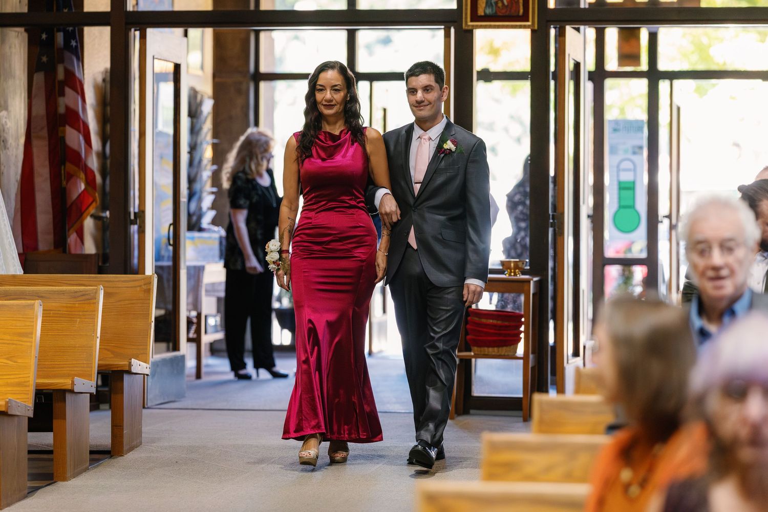 Formal event attendees walking through a sunlit hallway with wooden pillars.