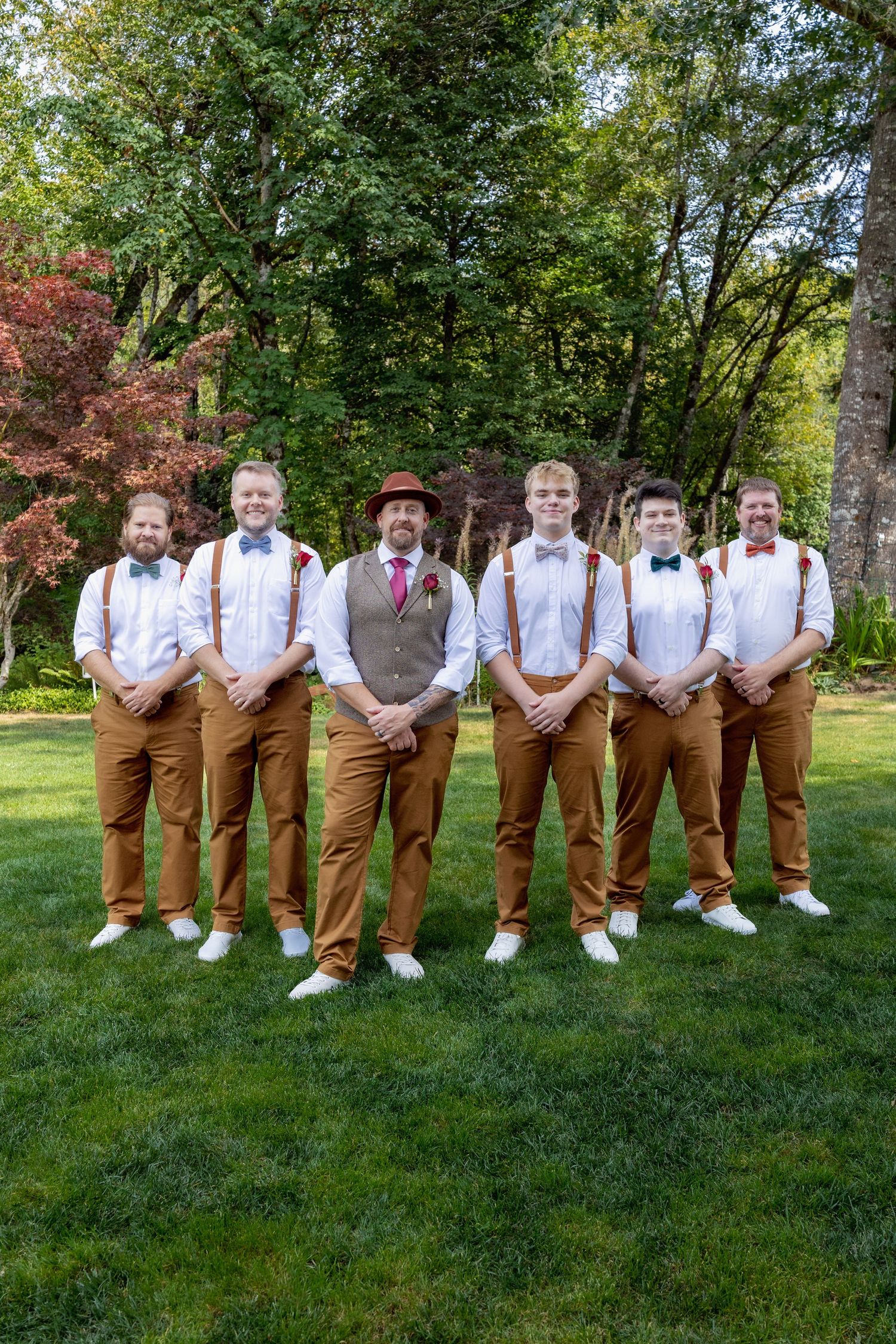 Groomsmen in matching brown pants and white shirts posing on grass with trees in background.