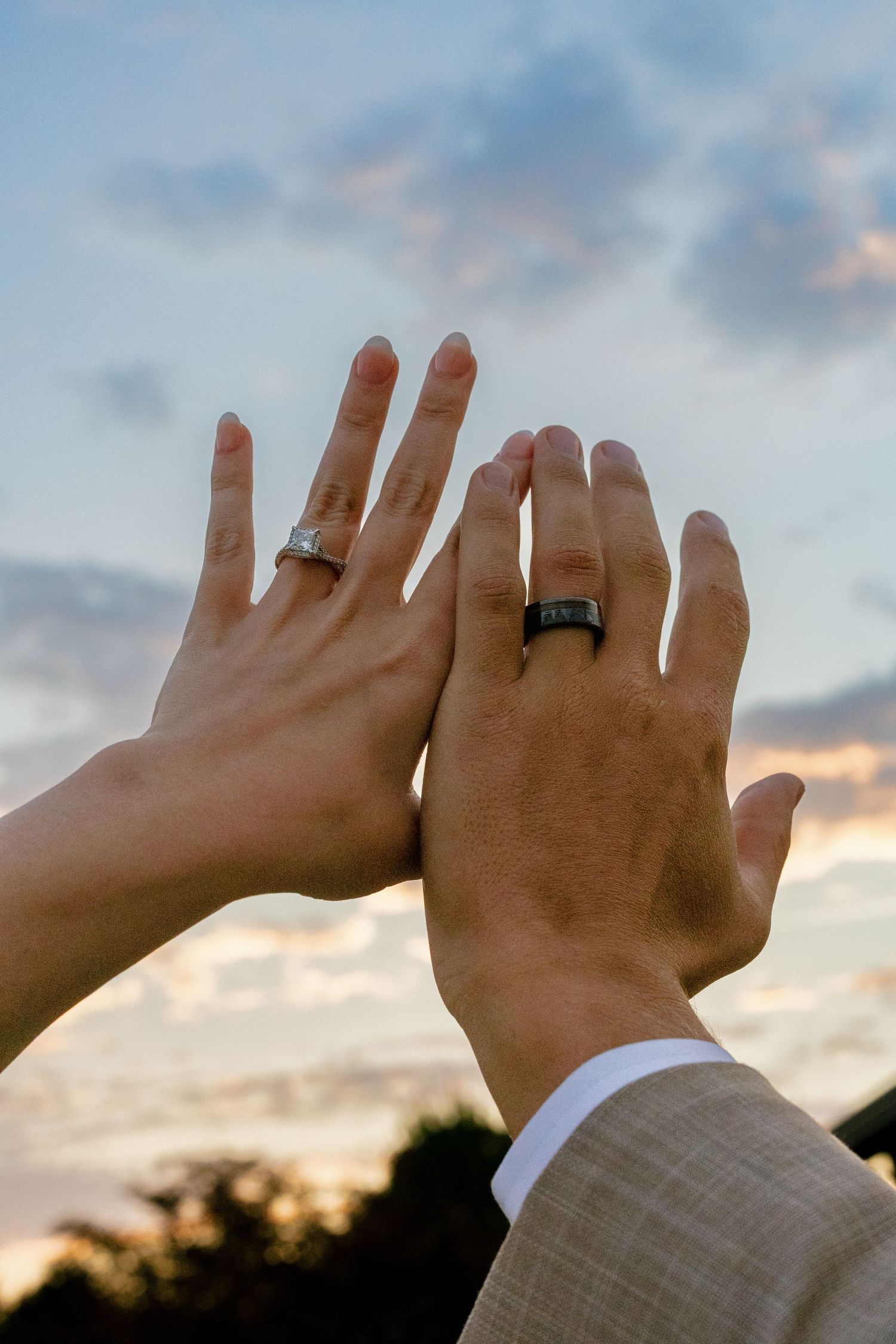 Two hands wearing wedding rings are raised against a sunset sky with clouds.