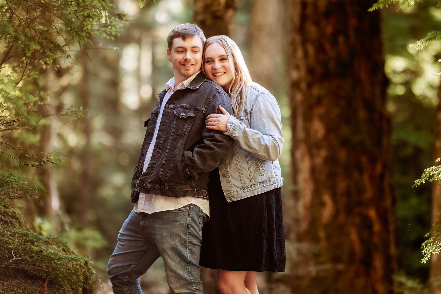 Couple embraces in romantic forest engagement photo session with sunlit trees in background.