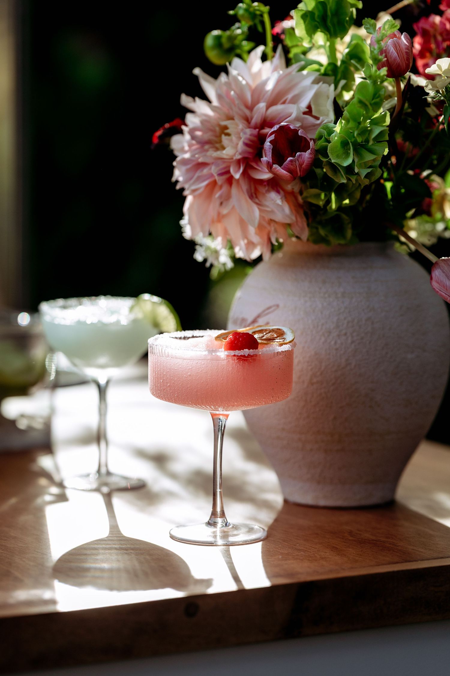 Pink cocktails in elegant coupe glasses sit beside pink dahlias in a vase with natural lighting.
