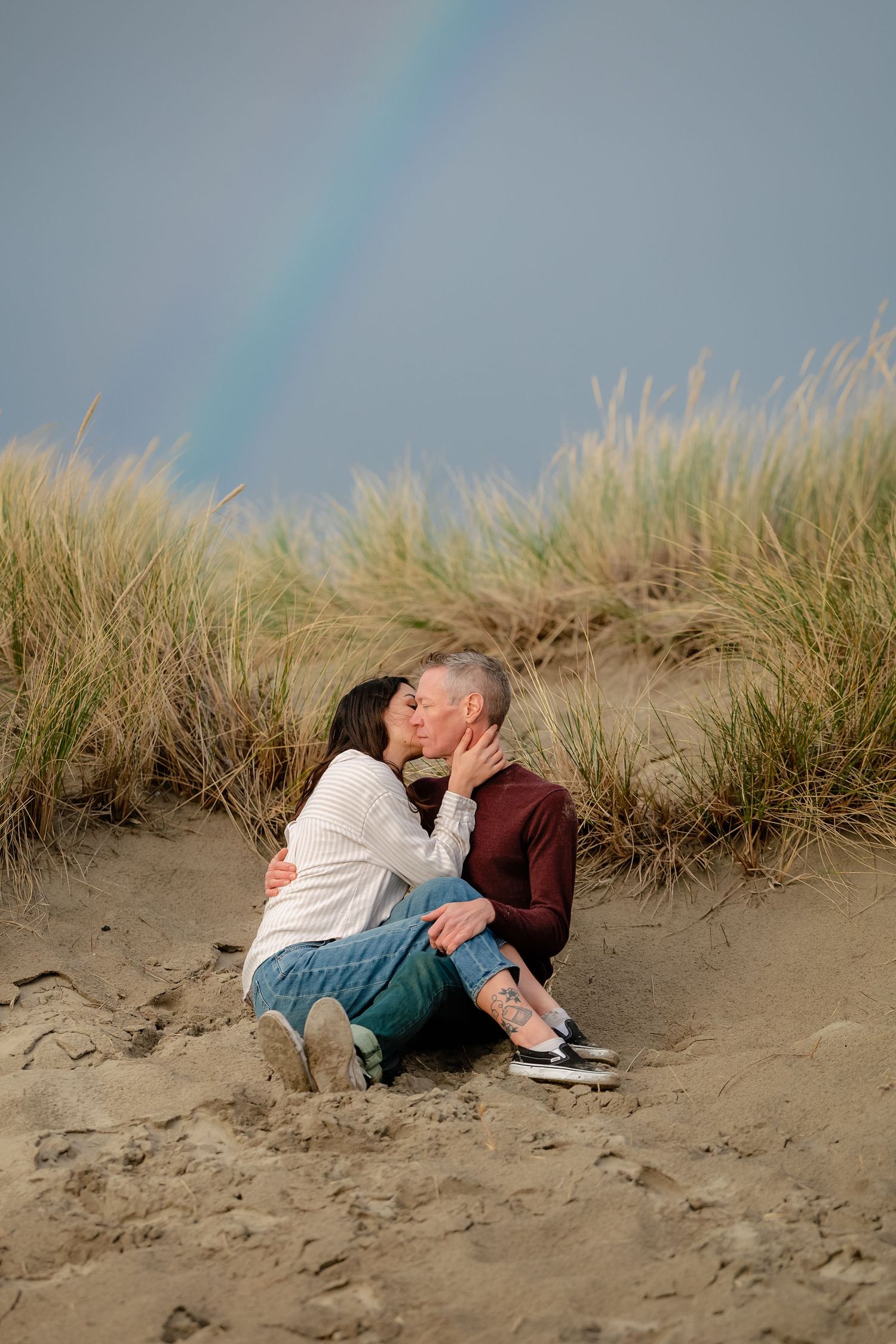 A romantic couple reclines together on sandy beach dunes with tall grass swaying in the background.